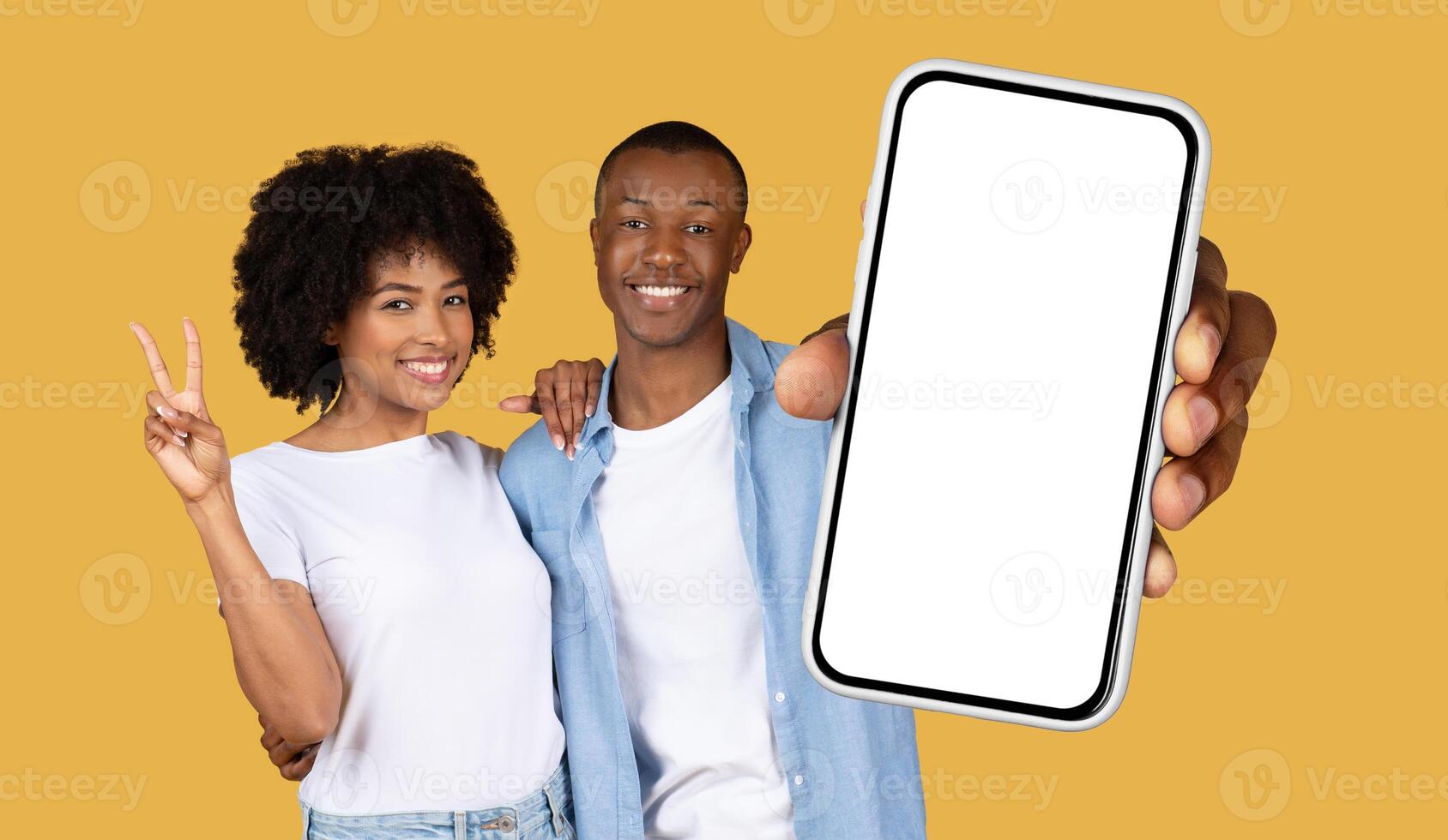 African American young, smiling couple standing in front of a yellow background. The man is holding a smartphone with a blank screen towards the camera, while the woman is making a peace sign photo