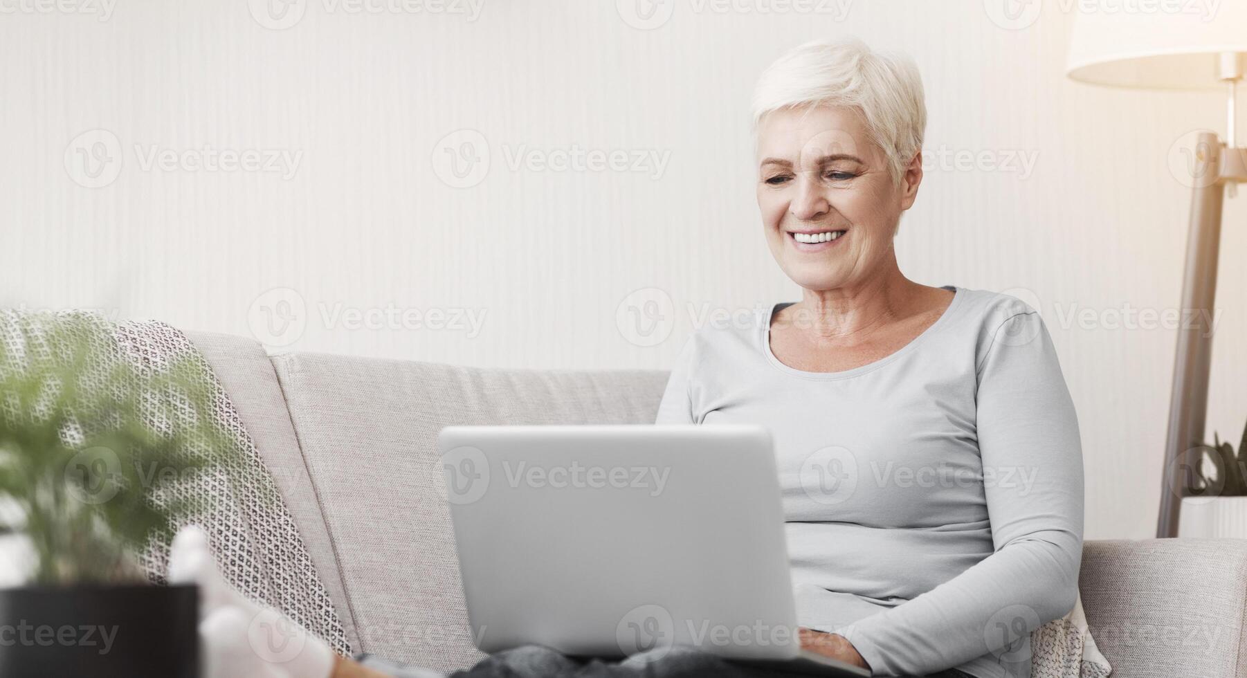 Smiling old woman working from home, typing email on laptop, sitting on sofa, copy space photo