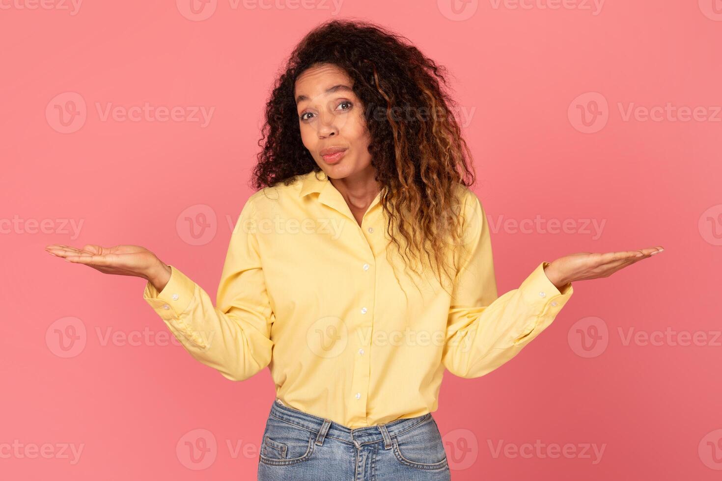 A young African American woman with curly brown hair stands in front of a solid pink background. She has her arms out to the side, palms facing up, and is looking with questioning expression. photo