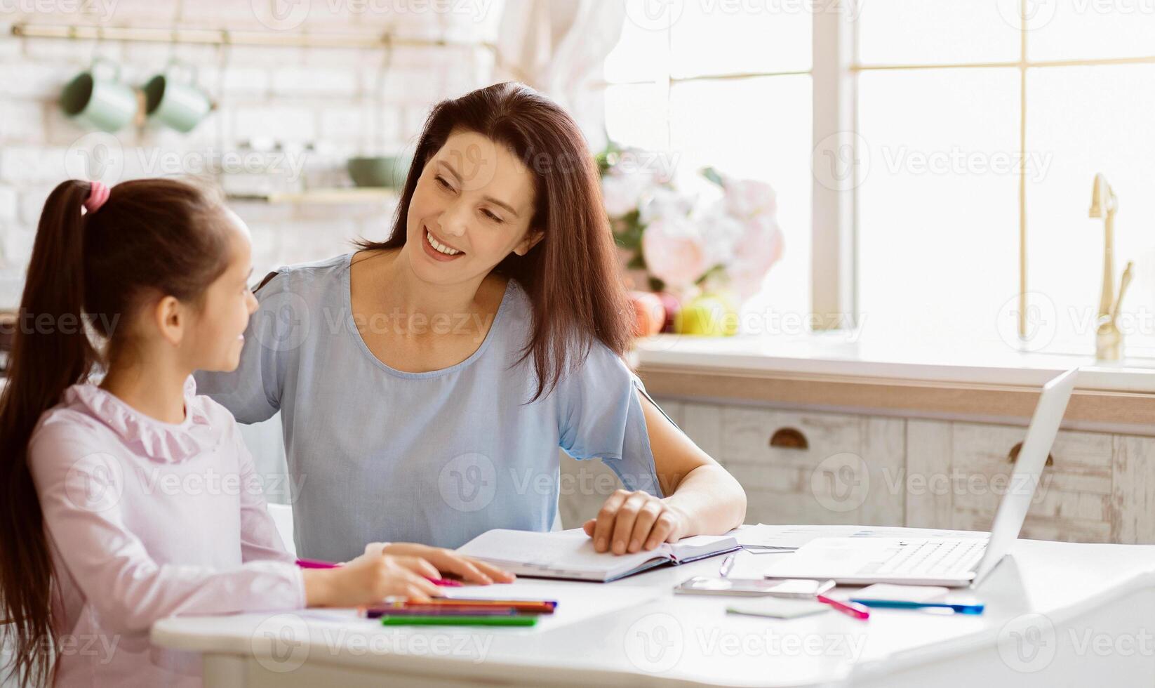 A mother and her young daughter are working together on homework at a table in their home. They are both smiling and look happy, copy space photo