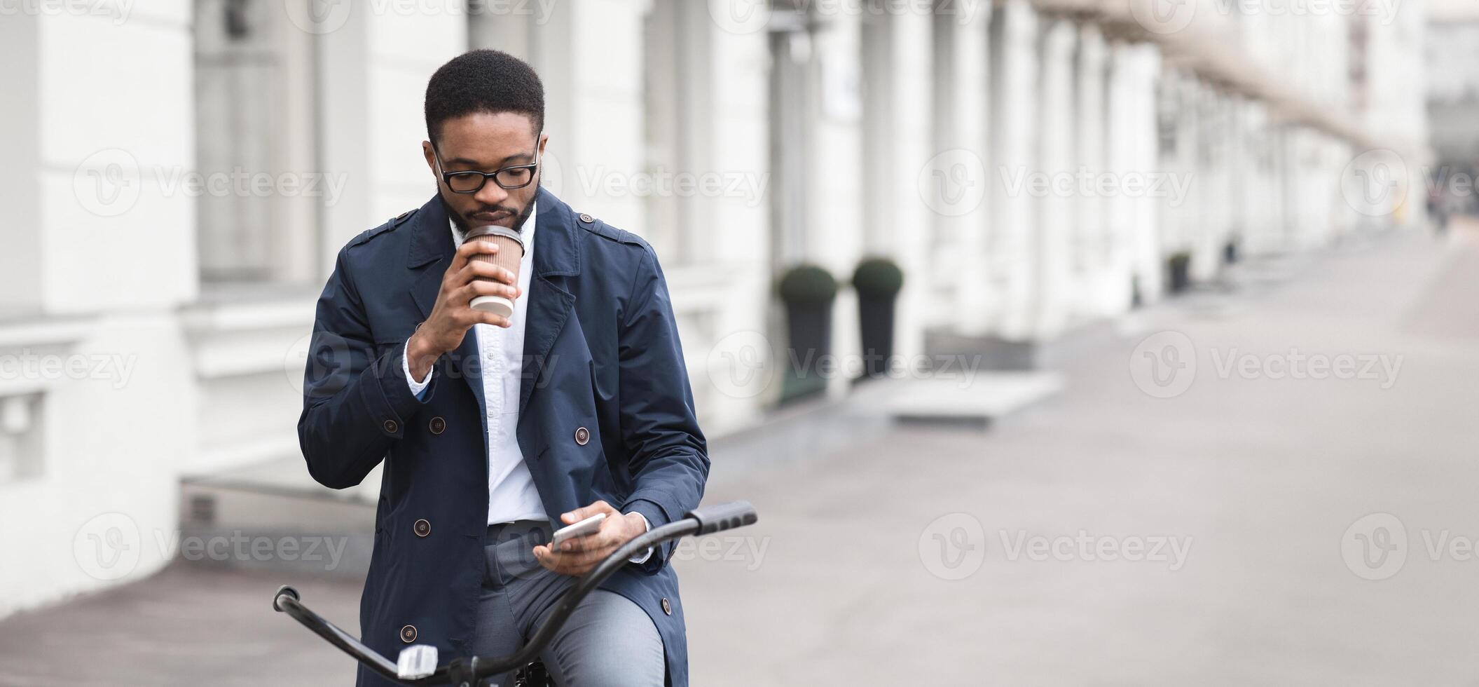 Black man professional is riding a bicycle to the office, carrying coffee and checking phone. The scene is modern and busy, capturing the essence of multitasking and an active start to the day. photo