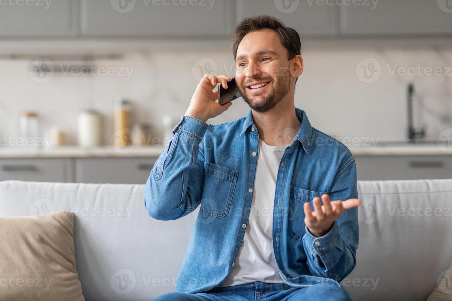 A man with a beard is sitting on a white couch in his home while talking on his phone. He is wearing a blue denim shirt and jeans. He is smiling and gesturing with his hand as he speaks. photo