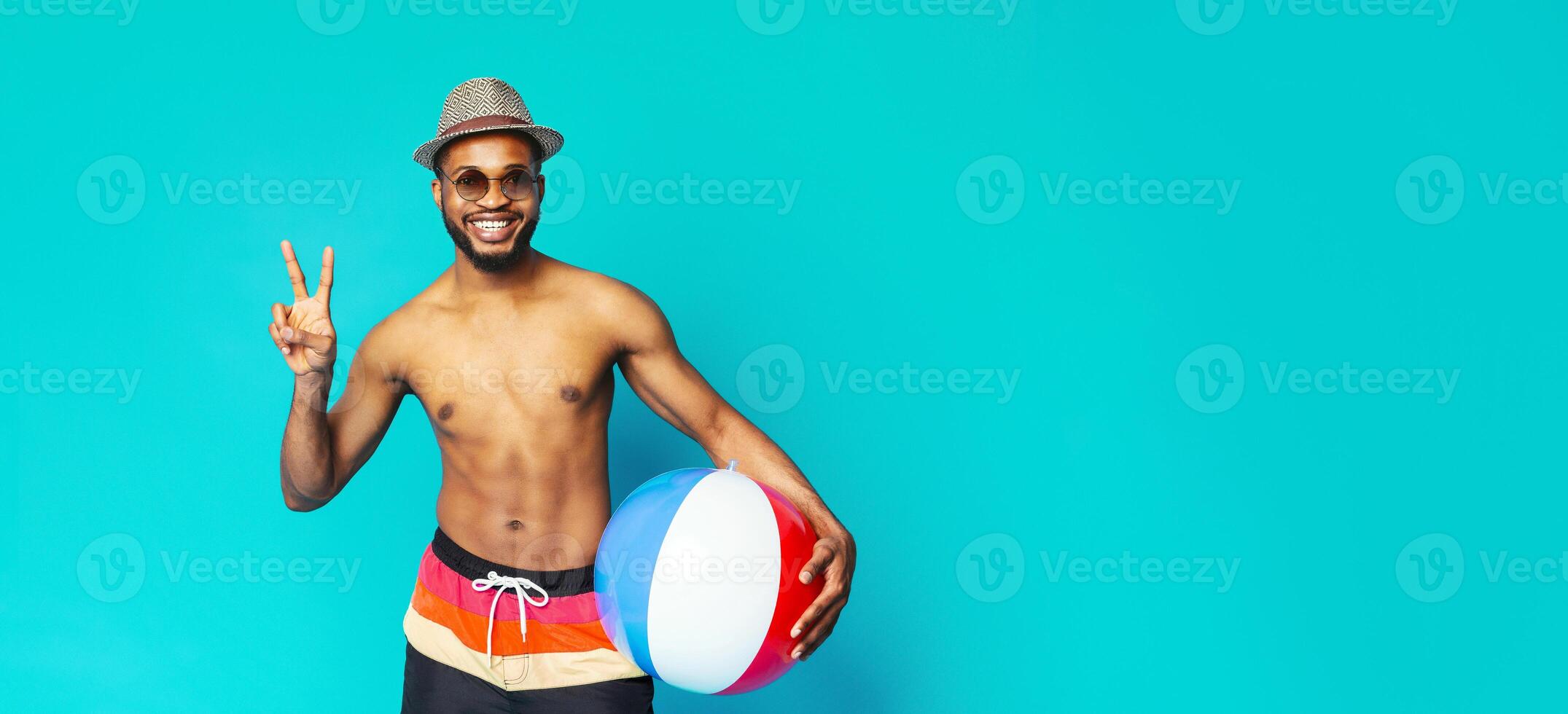 A smiling black man with a beard wearing sunglasses and a hat holds a beach ball while standing in front of a solid blue background. He is wearing colorful swim trunks and making a peace sign photo