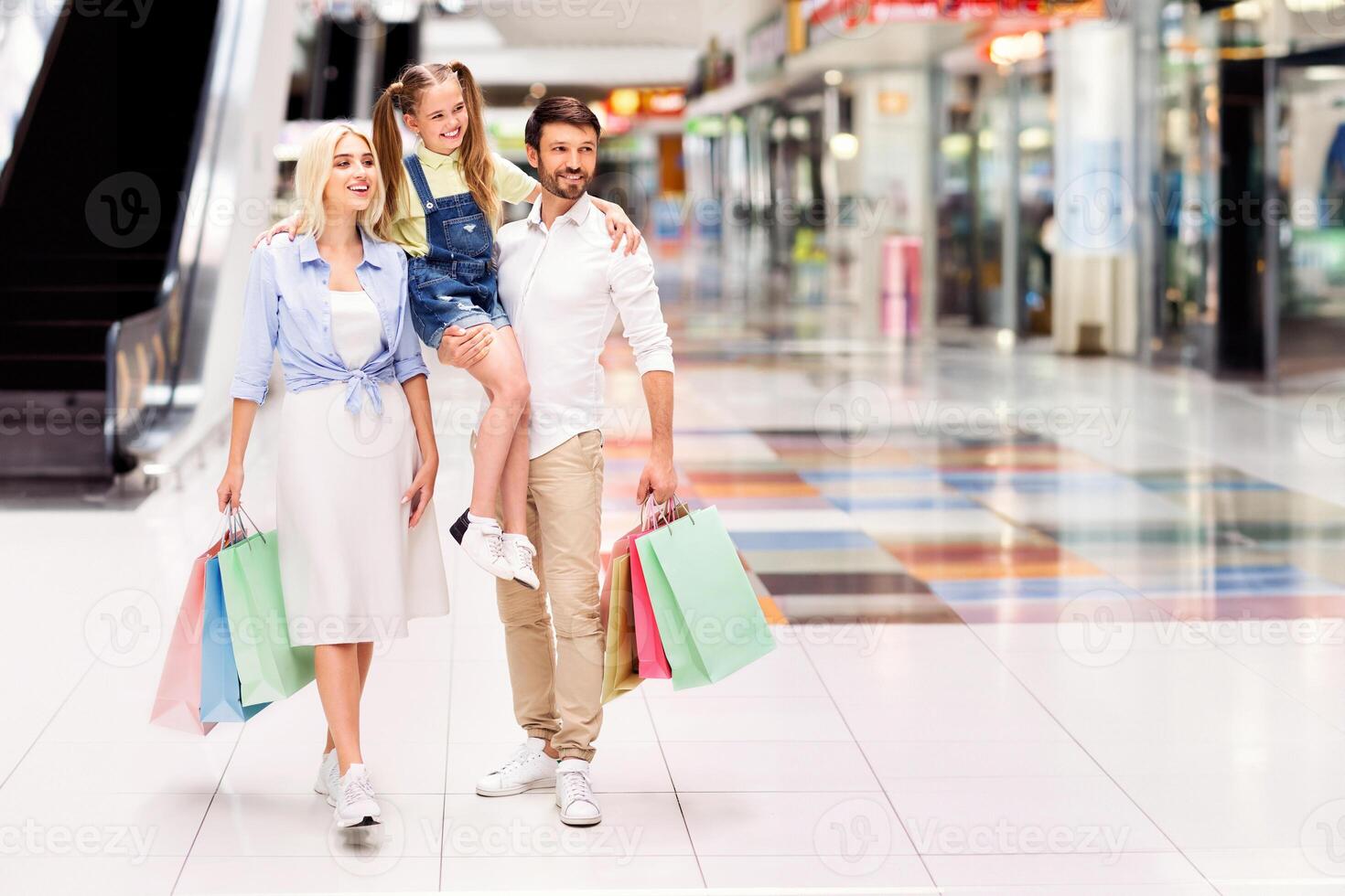 A family of three, a man, a woman, and a young girl, are walking through a modern shopping mall. They are carrying colorful shopping bags and smiling, copy space photo