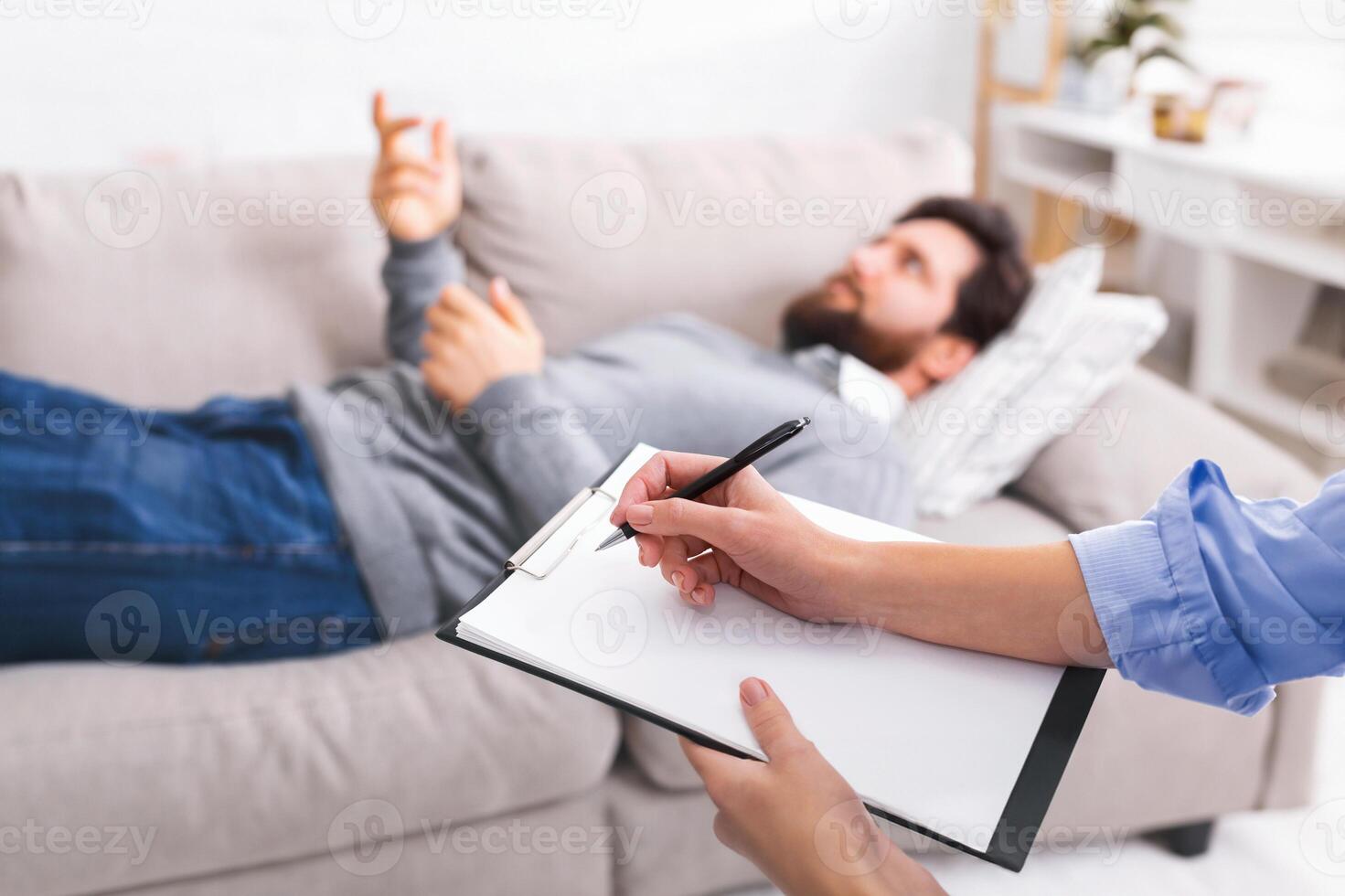 A man is lying on a couch, gesturing with his hand as he speaks, while engaged in a counseling session. A therapist, partially visible in the foreground, is attentively taking notes on a clipboard. photo