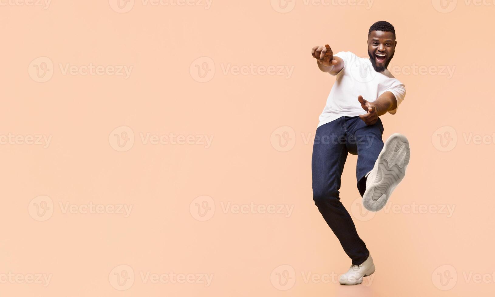 A young Black man in a white t-shirt and blue jeans is dancing in front of a peach-colored background. He has a wide smile on his face and is lifting his right leg, copy space photo