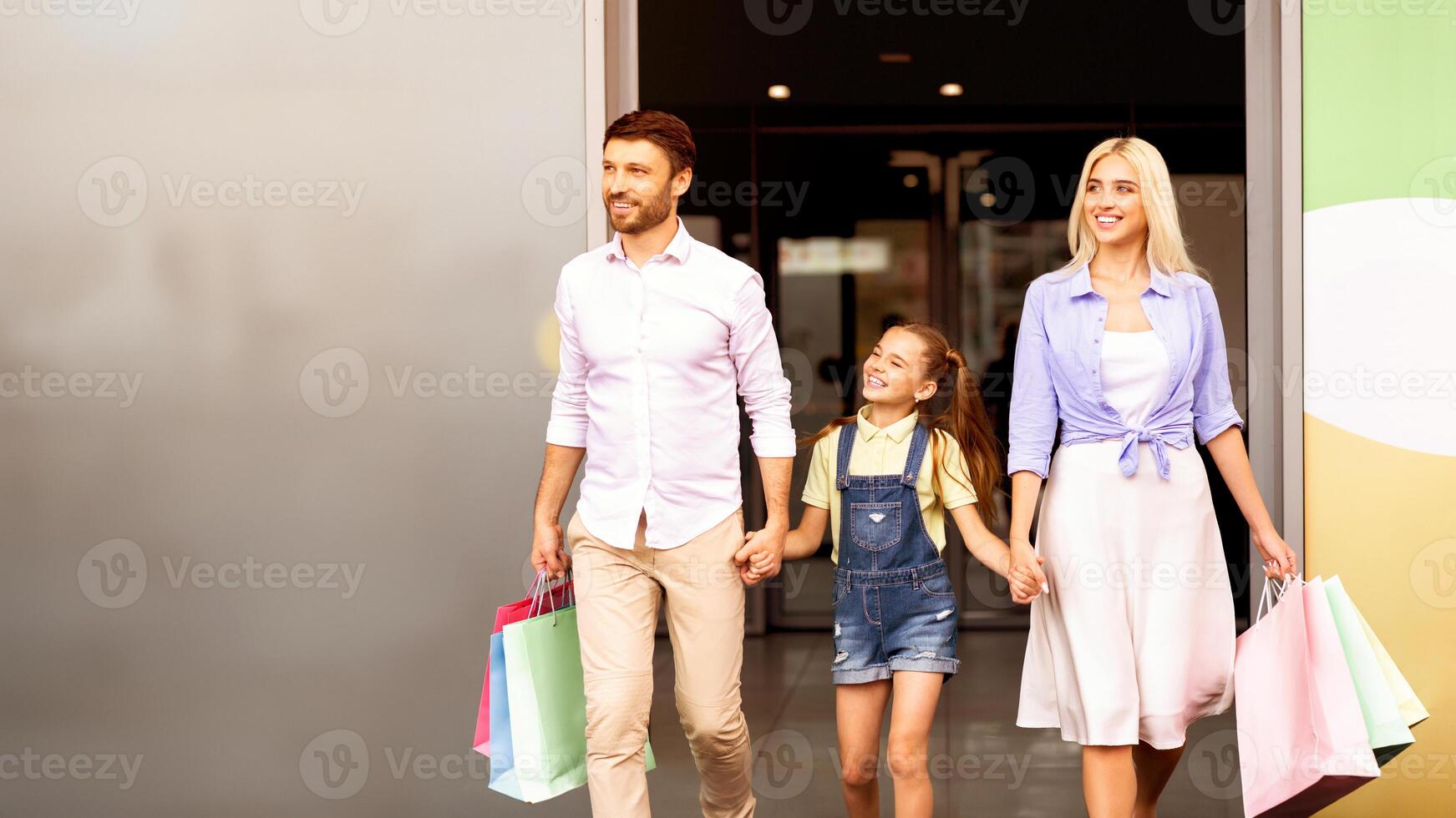 A family, consisting of a father, mother, and young daughter, are walking out of a shopping center. They are holding hands and carrying colorful shopping bags, copy space photo