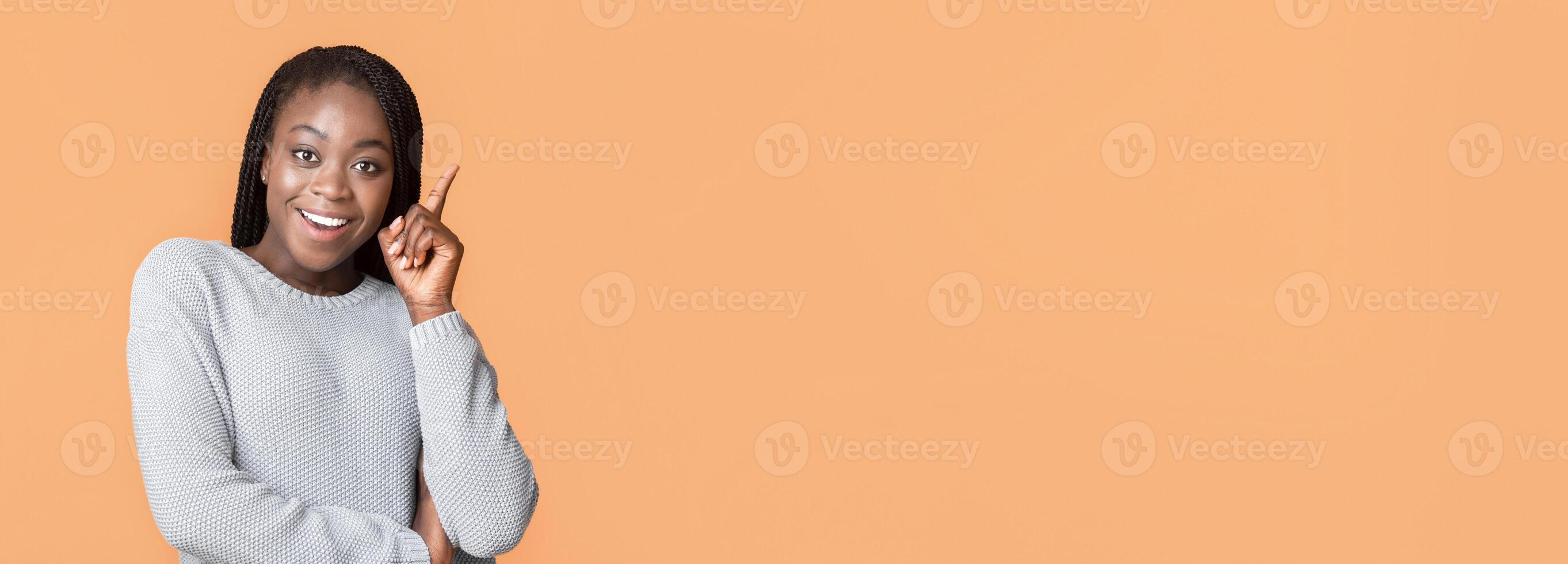 A cheerful young African American woman with braided hair stands in front of a vibrant orange backdrop. She wears a light gray sweater and confidently points up in eureka gesture photo