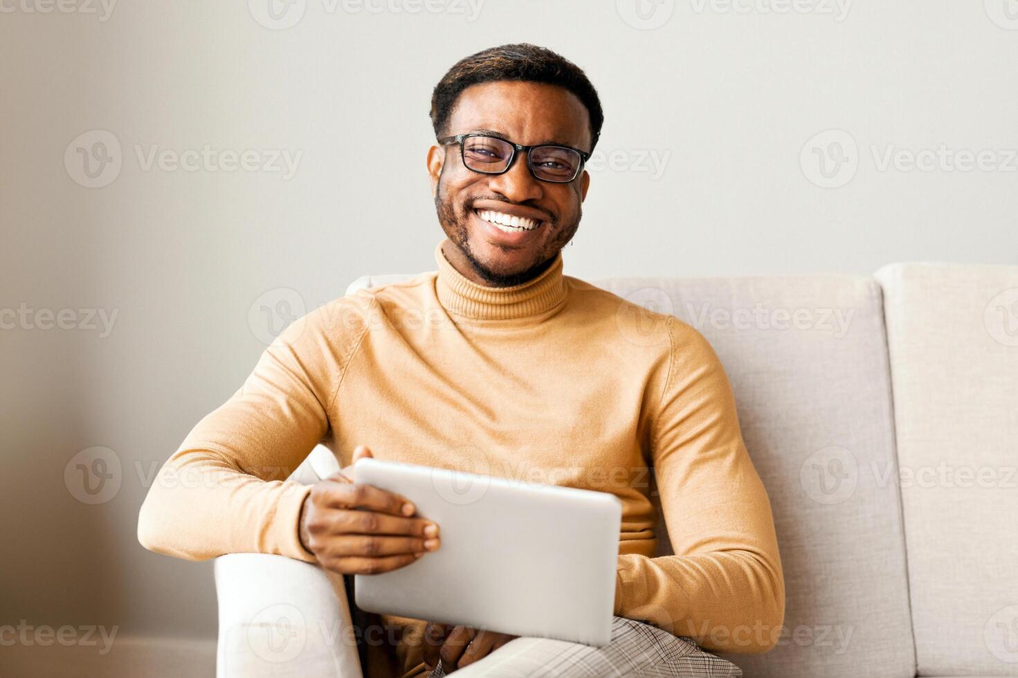 Young Afro Man Holding Digital Tablet Computer And Smiling At Camera Sitting On Couch Indoor. Selective Focus photo