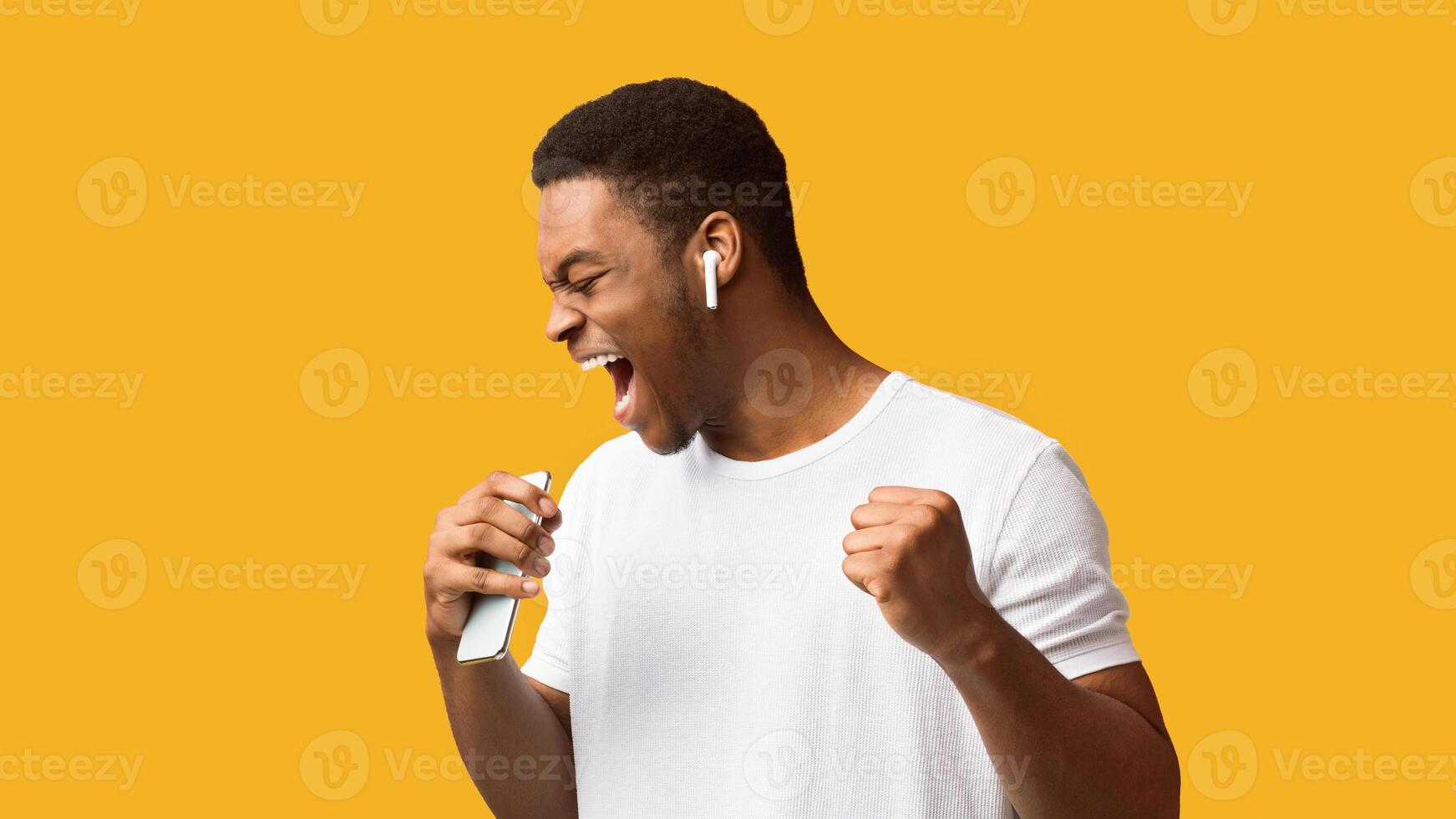 Joy Concept. Excited black man singing his favorite song, using smartphone as microphone, orange studio background photo