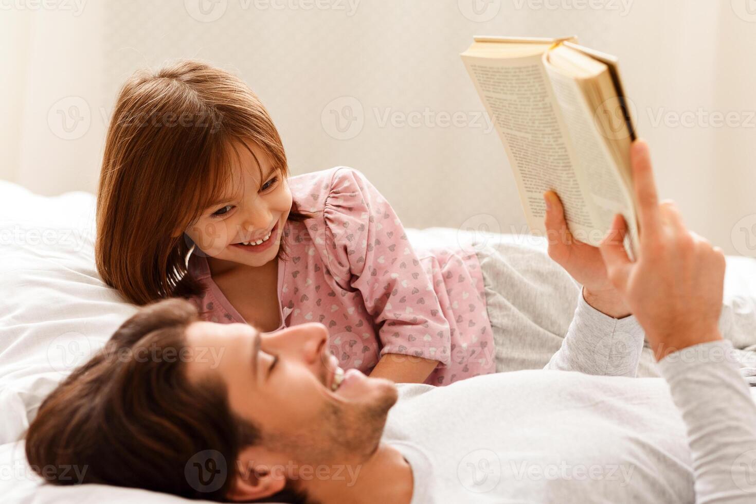 Young dad reading book to his little daughter, laying on bed together photo