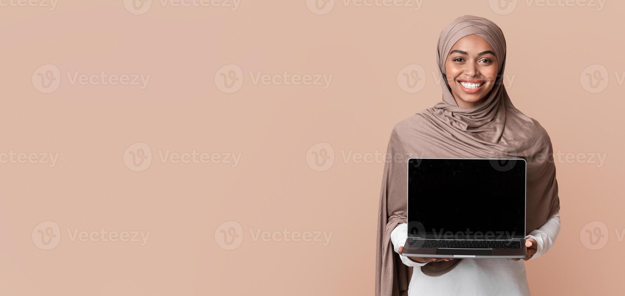 Cool Website. Smiling Black Muslim Girl In Hijab Holding Laptop Computer With Blank Black Screen For Mockup, Standing Over Yellow Background photo