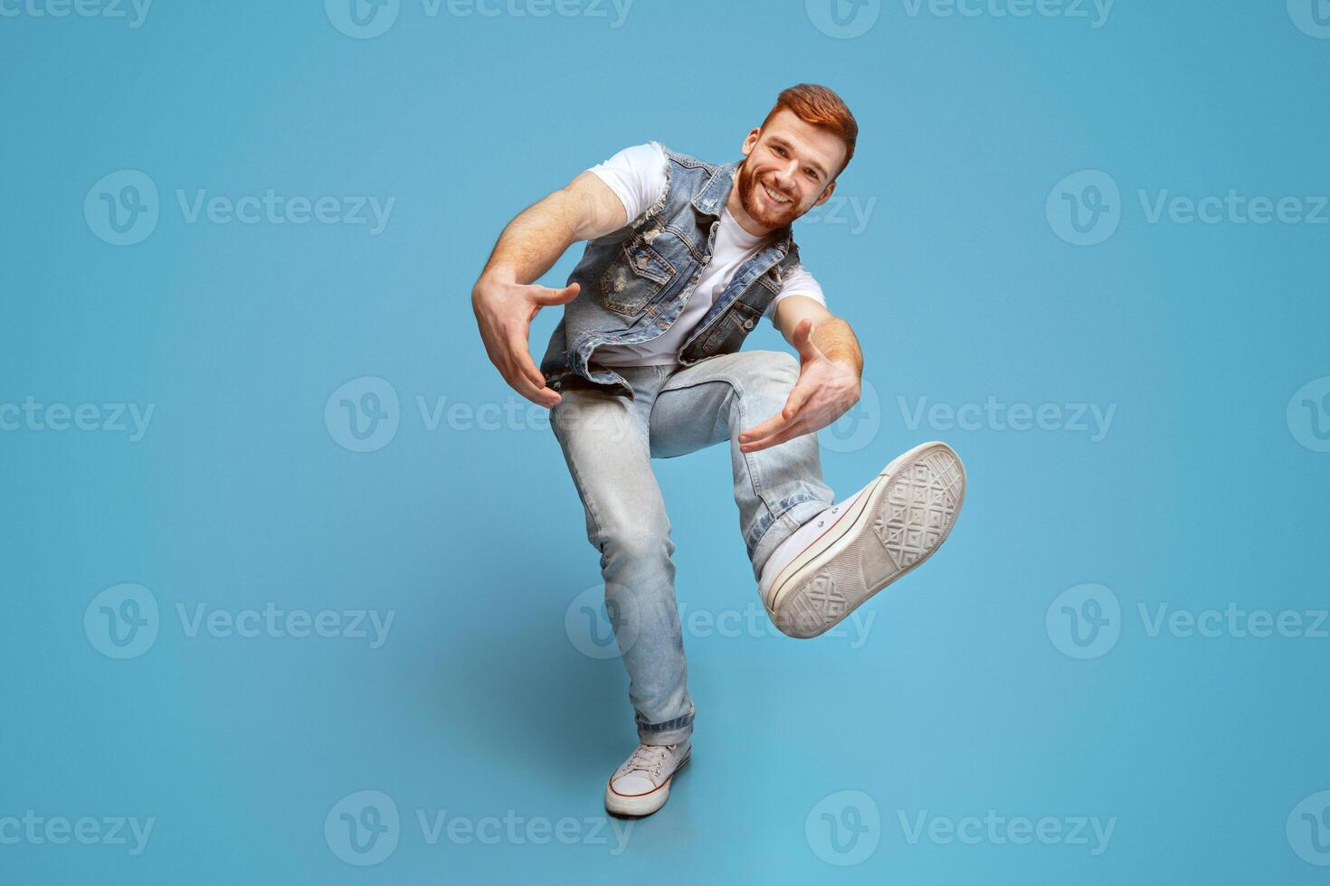 Come on. Smiling millennial caucasian guy fool around and showing his shoe sole at camera, full length, blue studio background photo