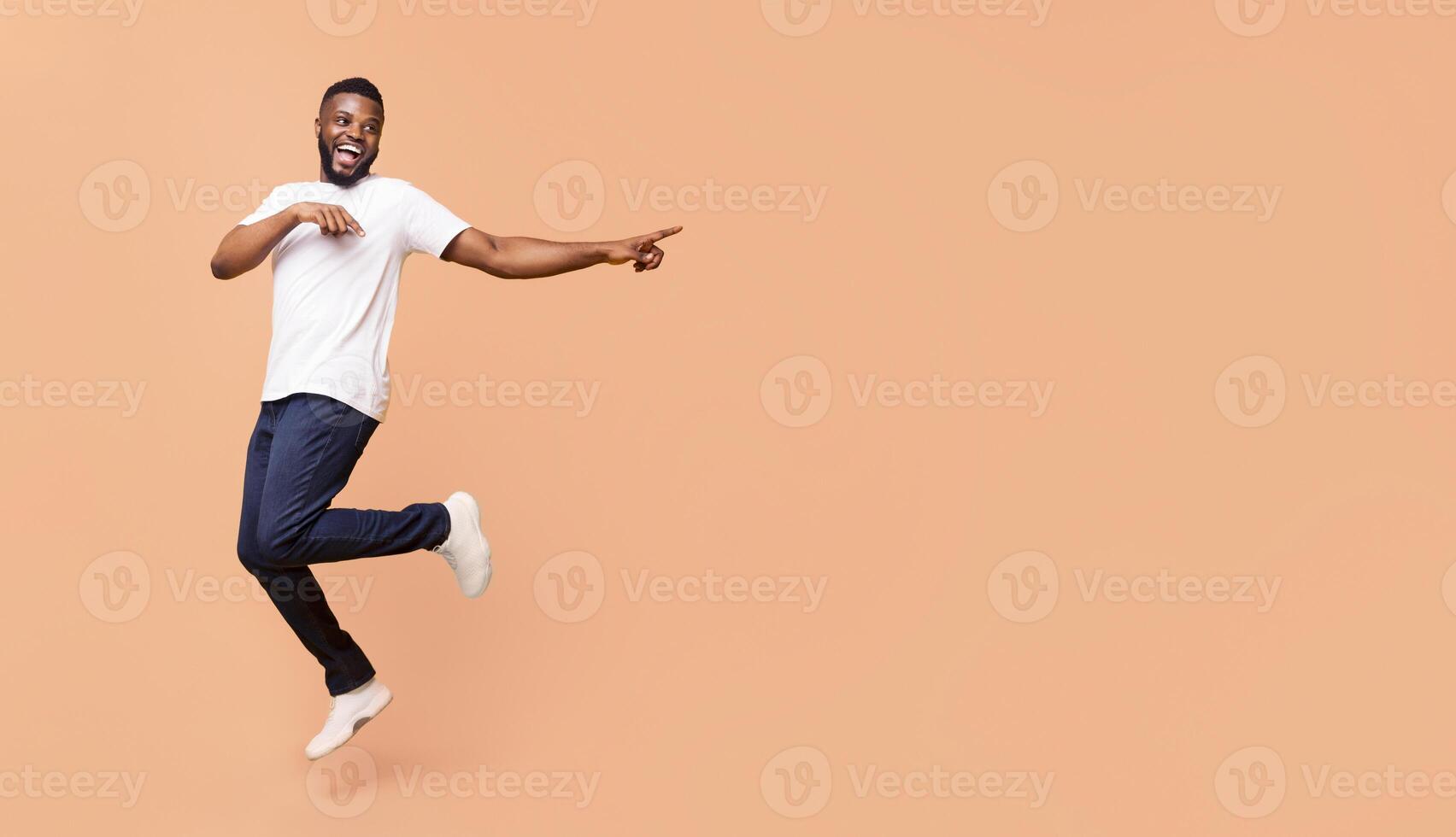 A happy African American young man is jumping in the air while pointing to the right with a big smile on his face. He is wearing a white t-shirt and dark blue jeans, panorama with copy space photo