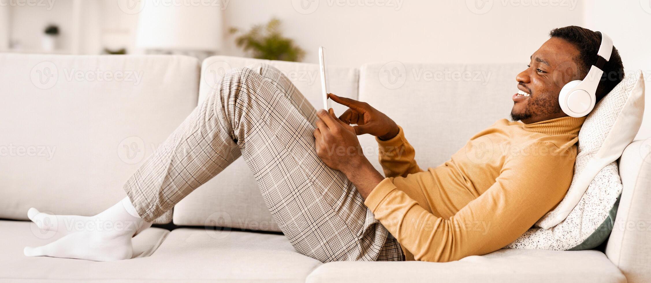 Gadget Lifestyle. Positive Afro Man In Earphones Using Tablet Computer Relaxing Lying On Sofa At Home photo