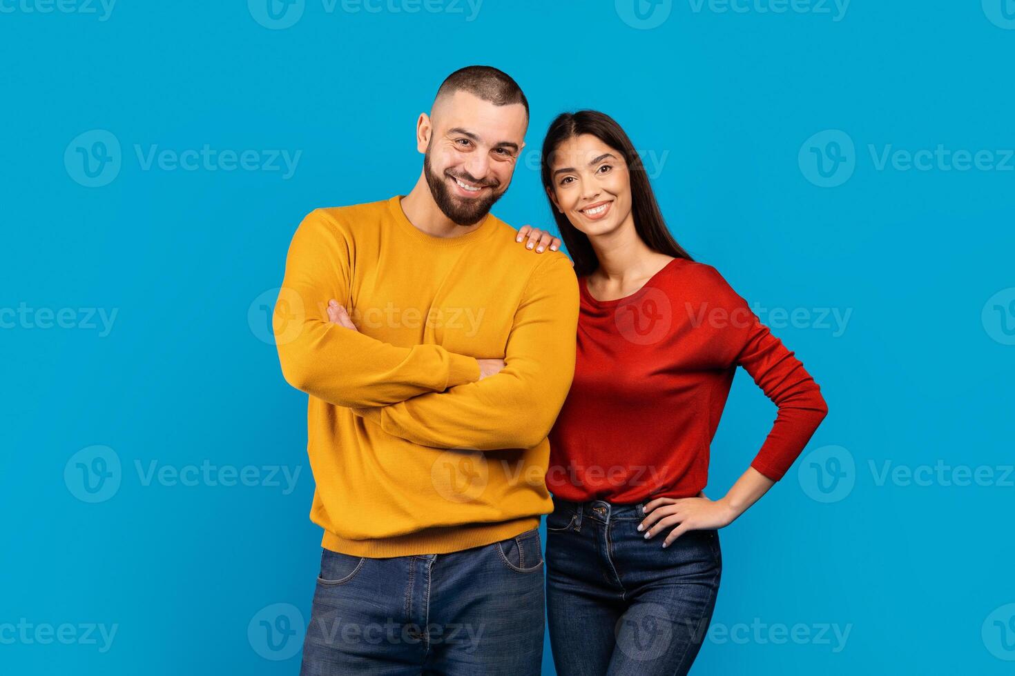 A man and woman standing together, smiling, and posing in front of the camera. They are dressed in casual attire and appear relaxed, blue background photo