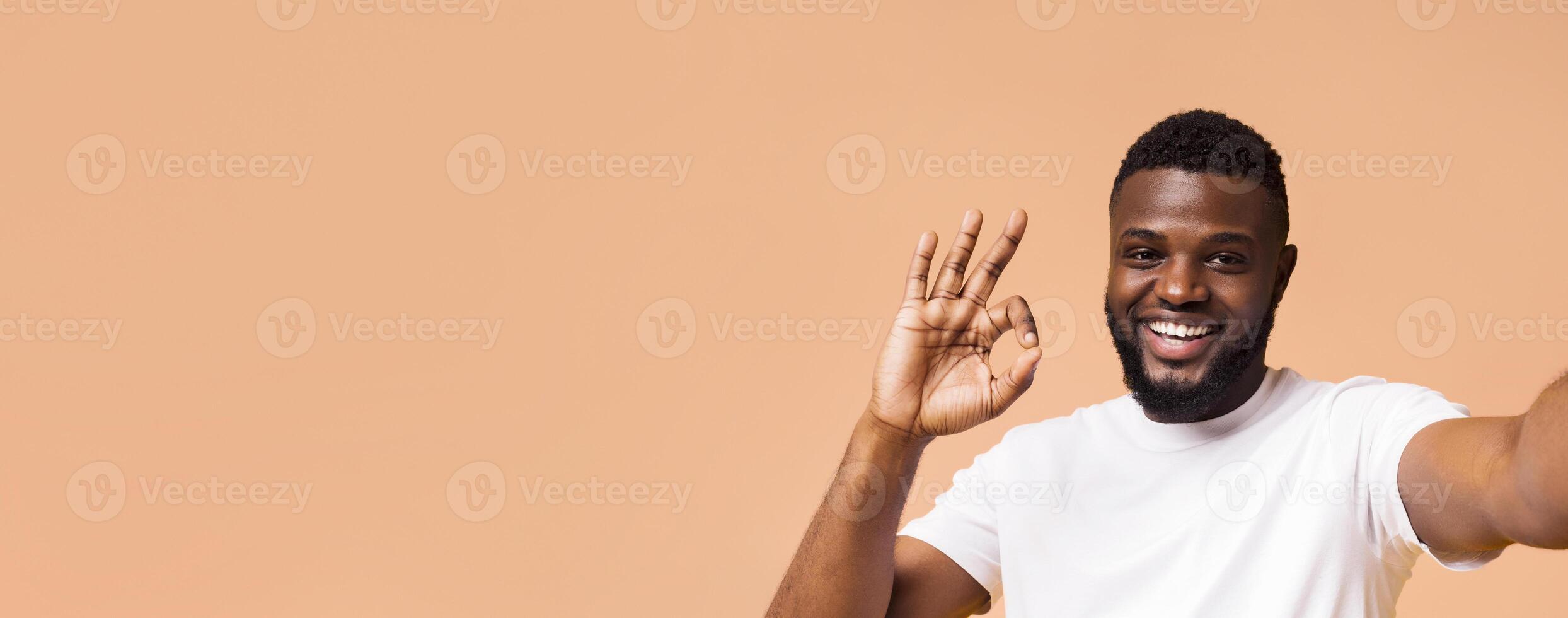 A smiling Black man with a beard and a white t-shirt is making the okay sign with his hand, looking directly at the camera. He is standing against a light brown background, copy space photo