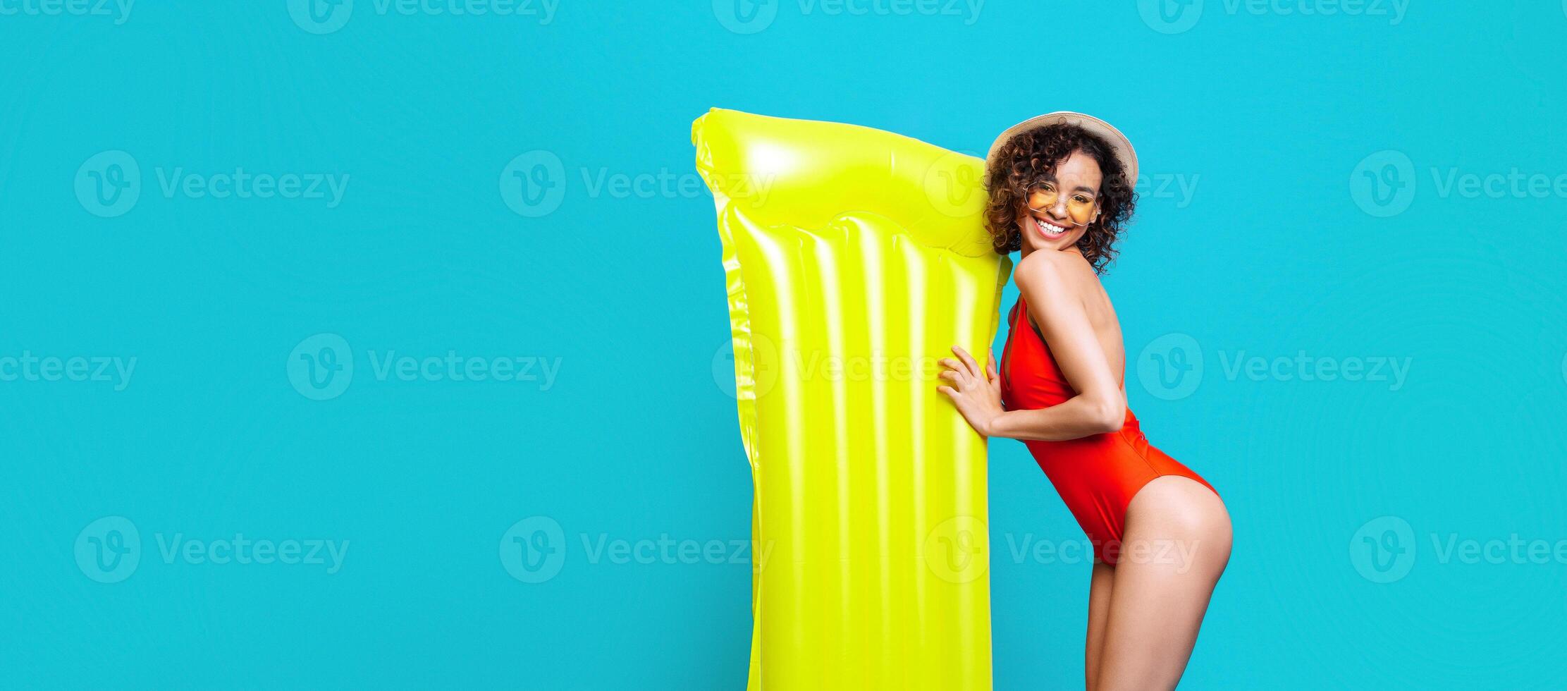 Black young woman in a red swimsuit and a straw hat poses with a yellow inflatable raft, smiling brightly against a turquoise backdrop. The scene embodies the carefree spirit of summer vacation. photo