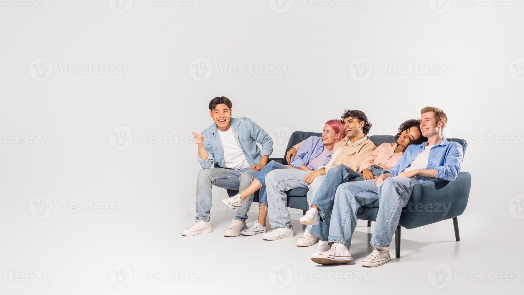 A group of five friends are enjoying each others company, laughing and smiling. The diverse group of young adults are casually dressed in denim and sitting on a couch in a light studio space. photo