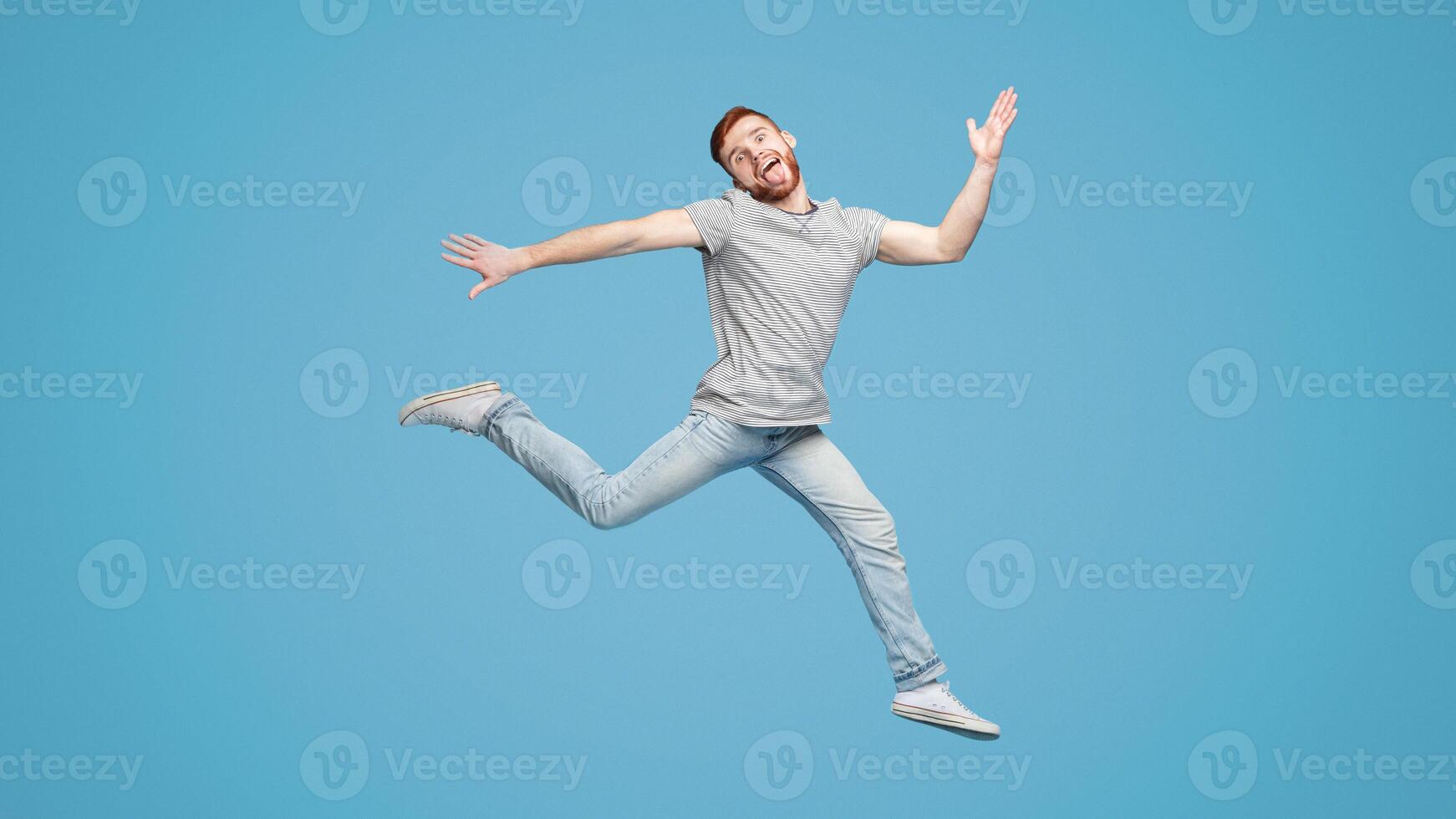 Funny millennial European guy jumping in air and fooling on blue studio background, full length, empty space photo