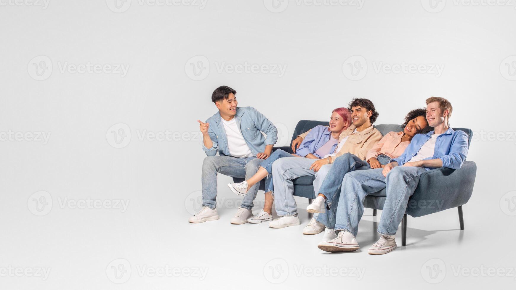A group of diverse friends enjoy each others company while sitting on a couch. One friend is seated slightly apart from the others, leaning forward with his arms crossed as he speaks, copy space photo