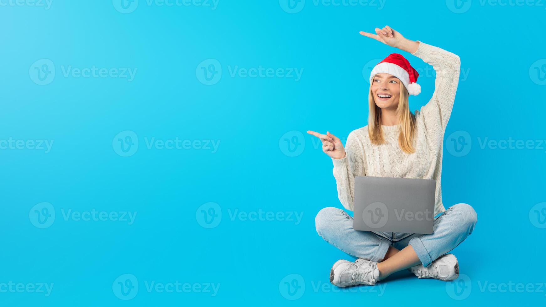 A woman is seated on the floor, wearing a Santa hat, and using a laptop. She appears to be focused on the screen as she types and scrolls through information, copy space photo