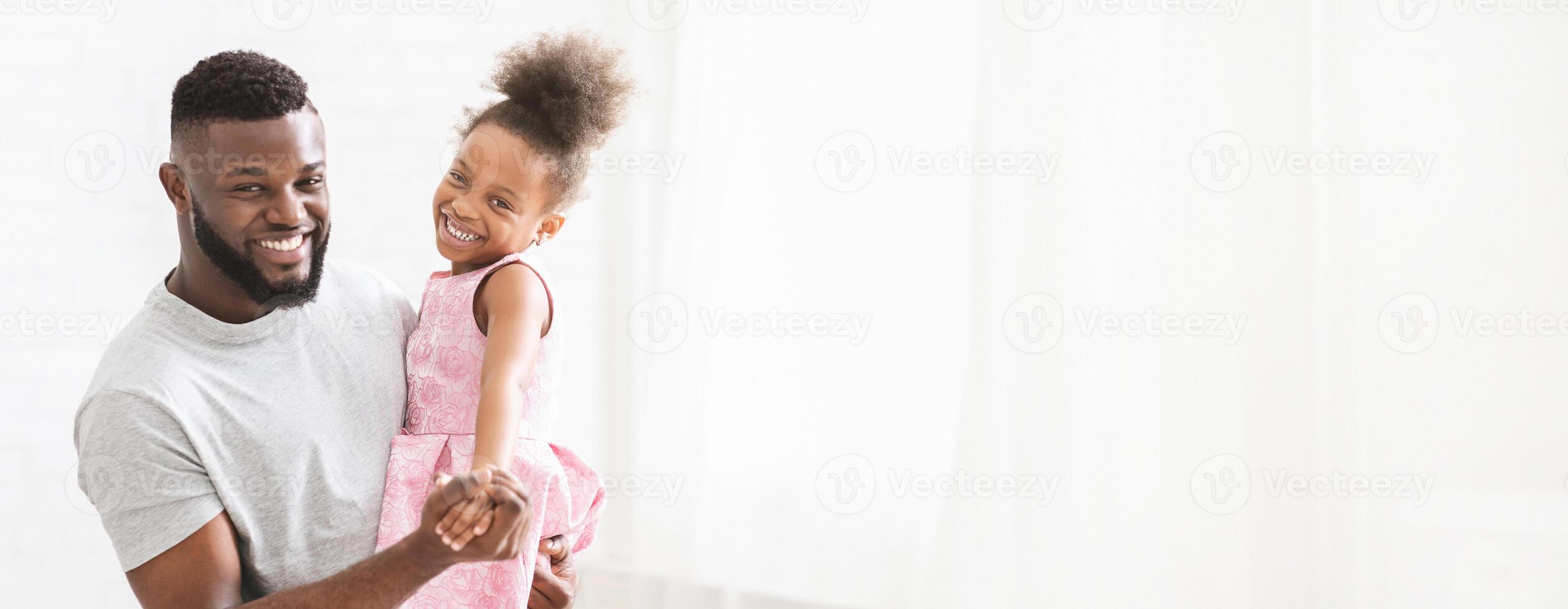 A joyful black father holds his smiling daughter in his arms as they dance playfully. Sunlight filters through the bright room, illuminating their happiness and creating a warm atmosphere. photo