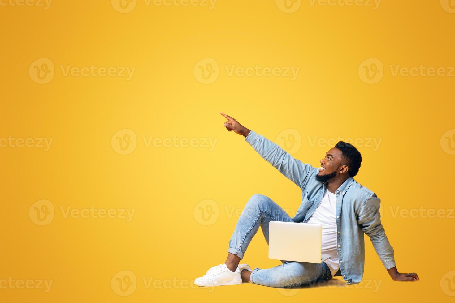 Look there. African american guy sitting on floor with laptop and pointing aside at copy space on yellow background photo