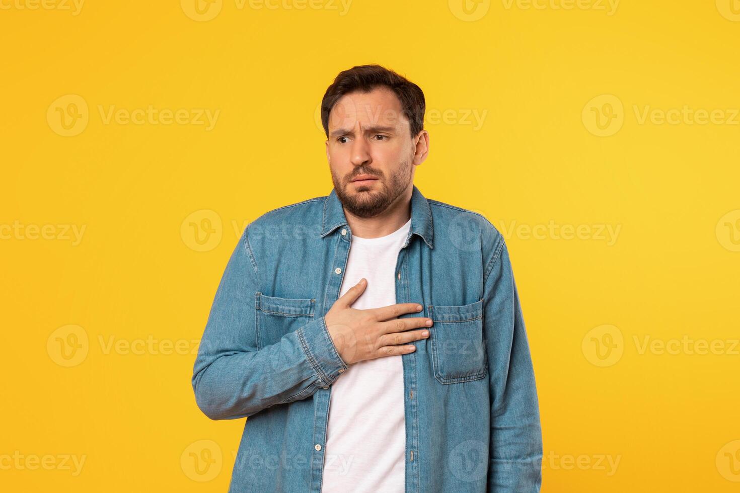 A man wearing a blue denim shirt and a white undershirt stands in front of a bright yellow background. He has a concerned expression on his face and is holding his hand to his chest. photo