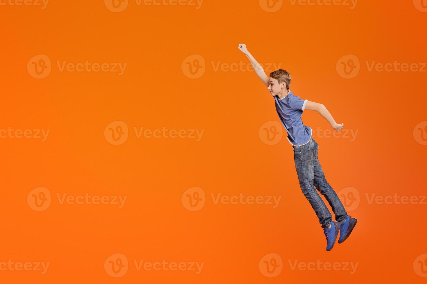 Little boy simulating flight, jumping with raised fist like superhero on orange background photo
