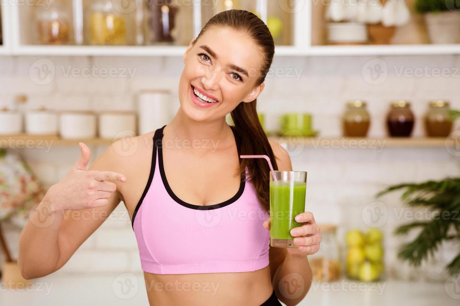 A woman in athletic wear smiles and holds a green smoothie in a glass with a straw. photo