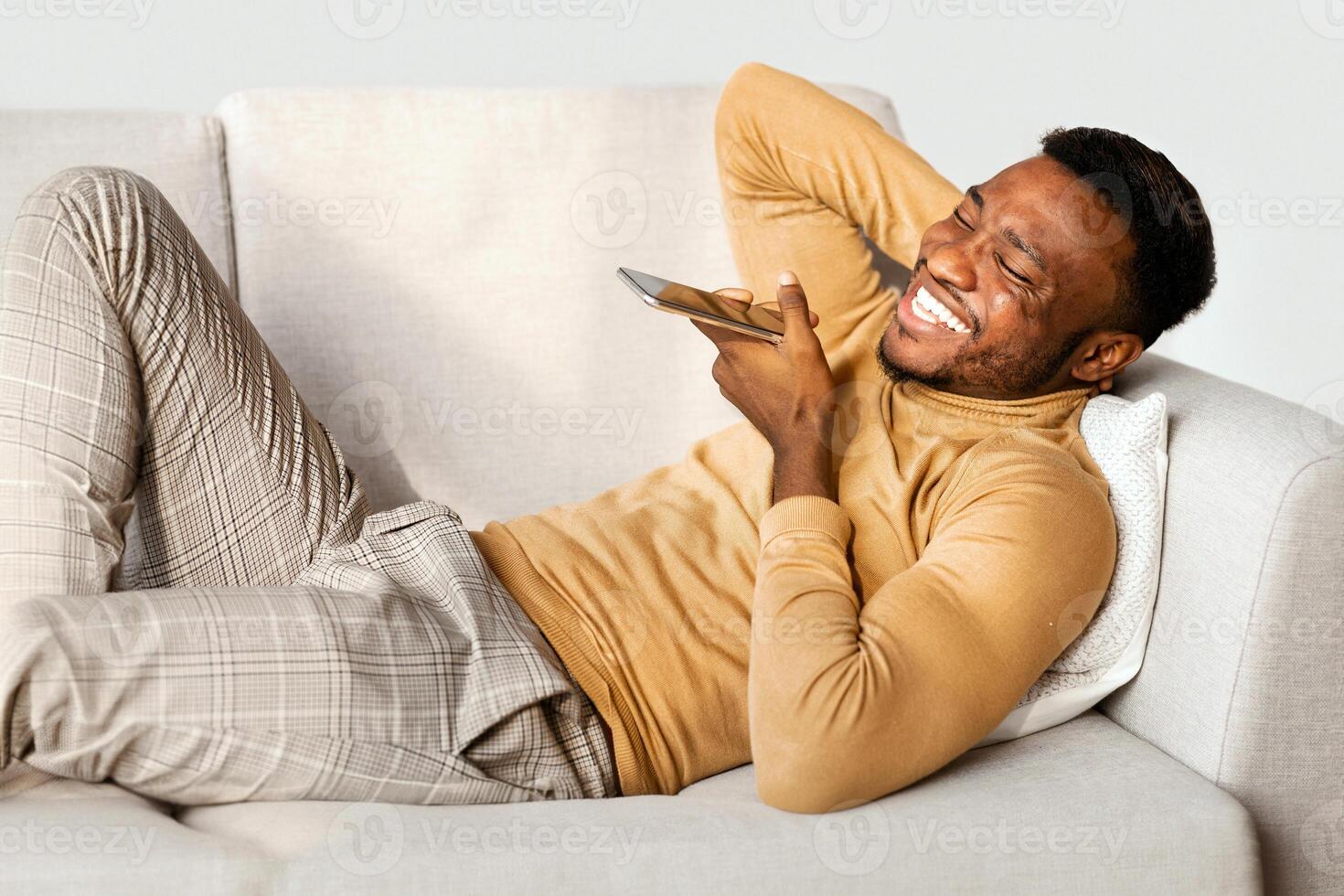 Positive Black Man Using Mobile Phone Voice Assistant Lying On Couch At Home. Modern Lifestyle photo
