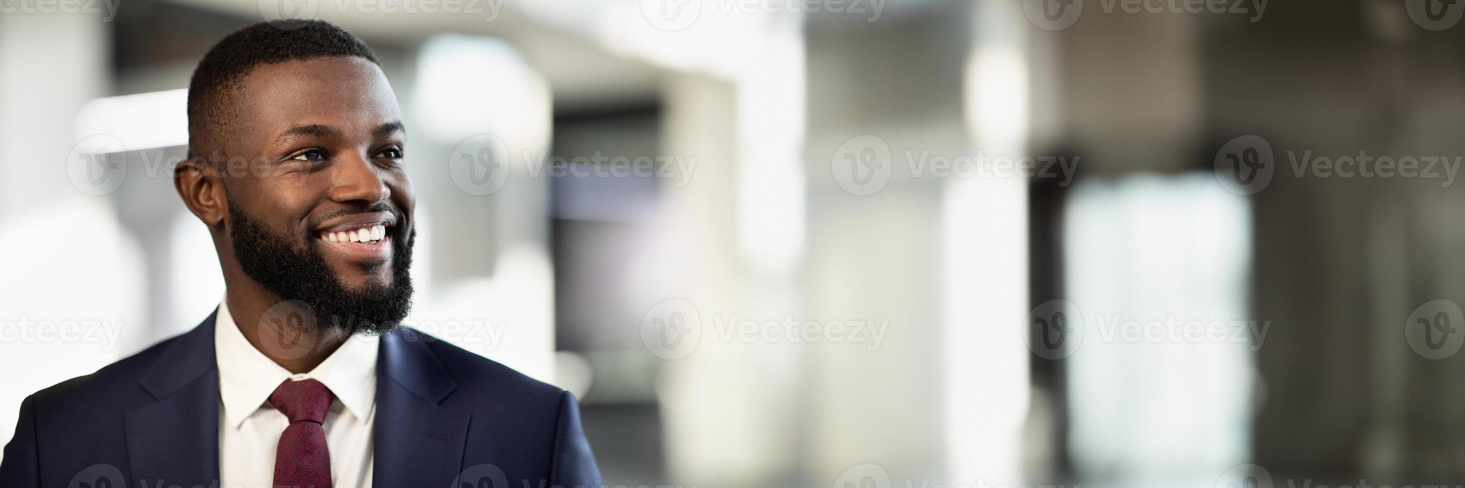 Closeup portrait of cheerful african american young businessman posing at office building, handsome black manager in suit and tie looking at copy space and smiling, panorama photo