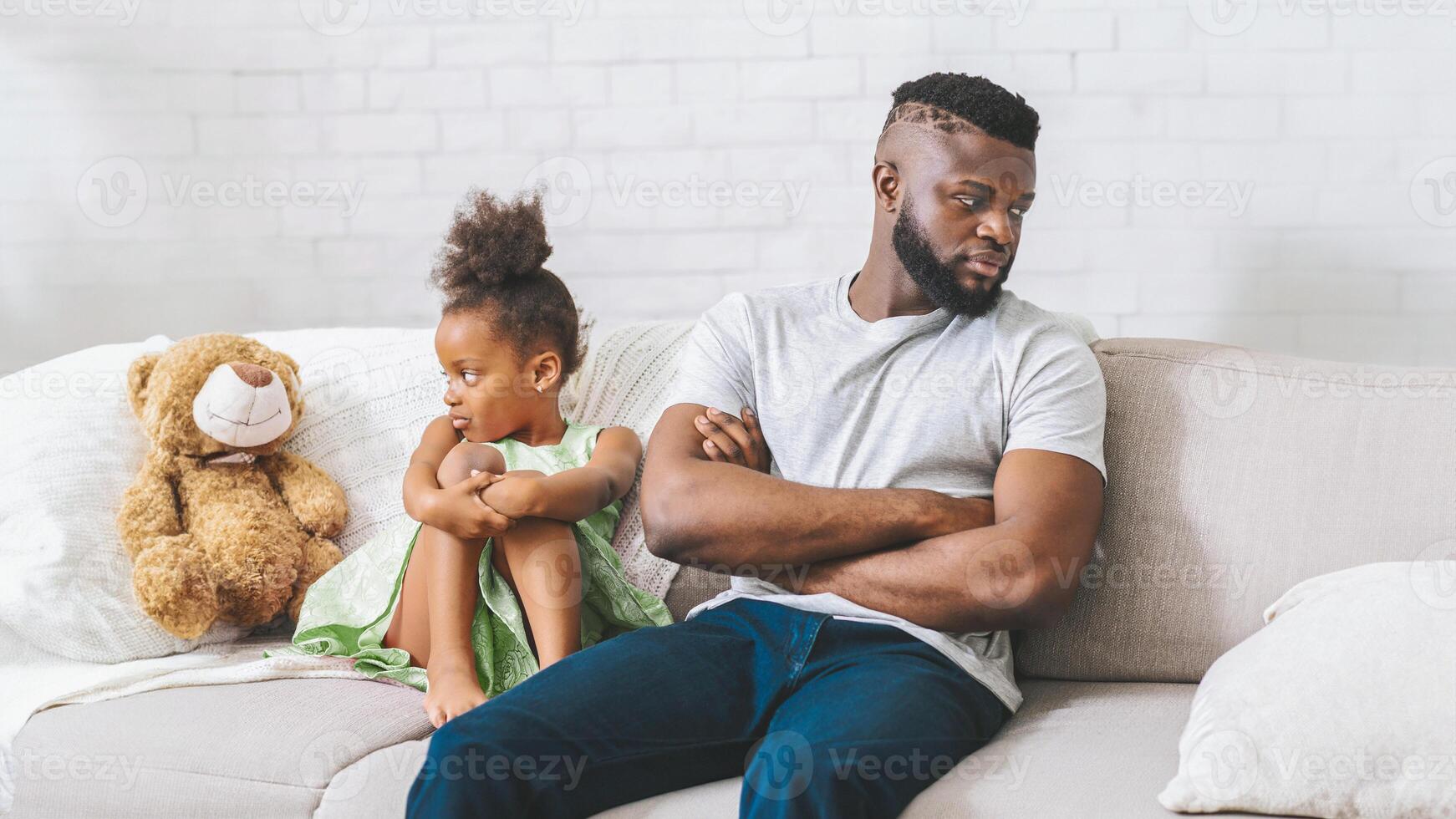 Black father and his young daughter are sitting on a couch, each looking away from the other. The father has his arms crossed and is looking down at the floor. The daughter is hugging her knees photo
