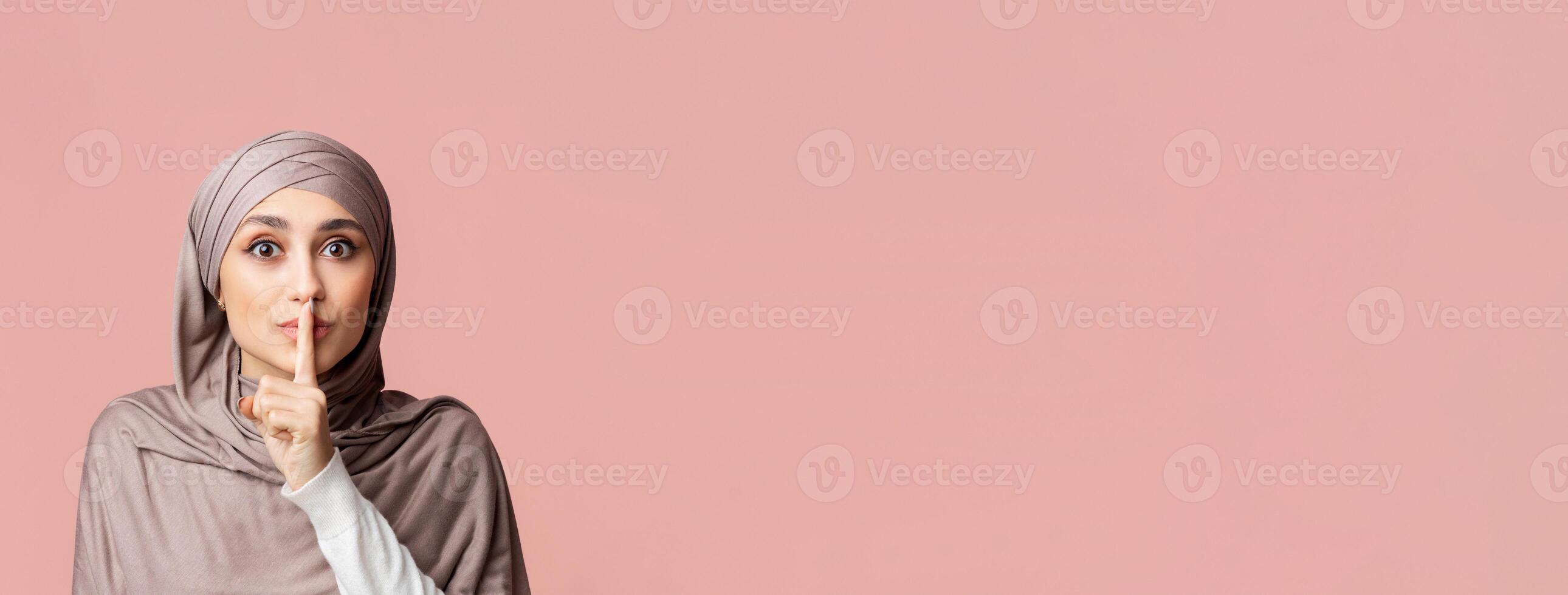 Secret Concept. Young arabic woman in hijab showing hush sign, putting finger on lips, gesturing keep silence over pink studio background, empty space photo