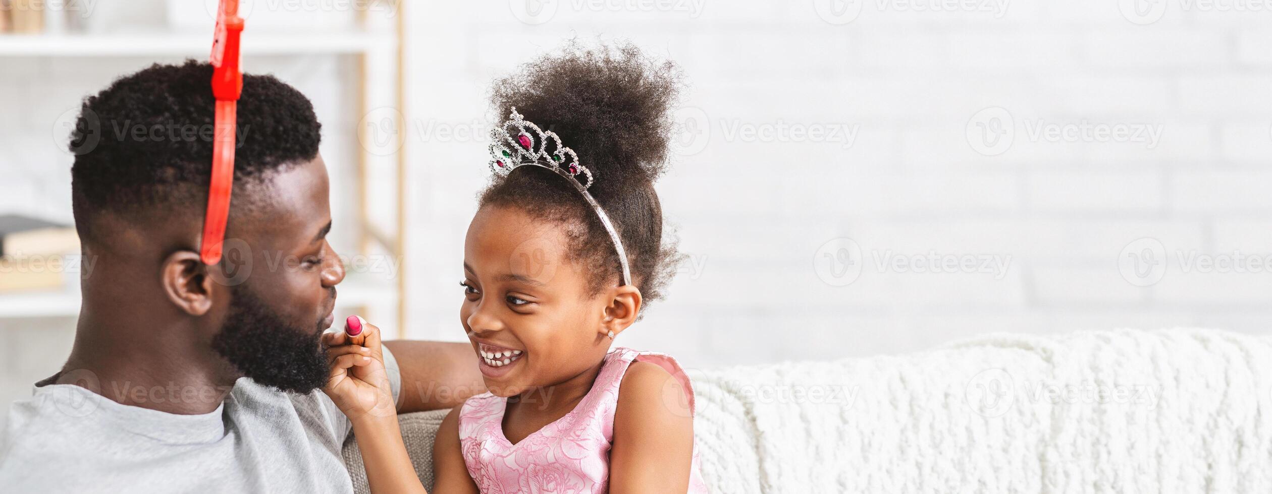 Black father and his young daughter share a playful moment on a cozy couch, both wearing costumes. The girl's laughter fills the room as she playfully touches her father's face, copy space photo