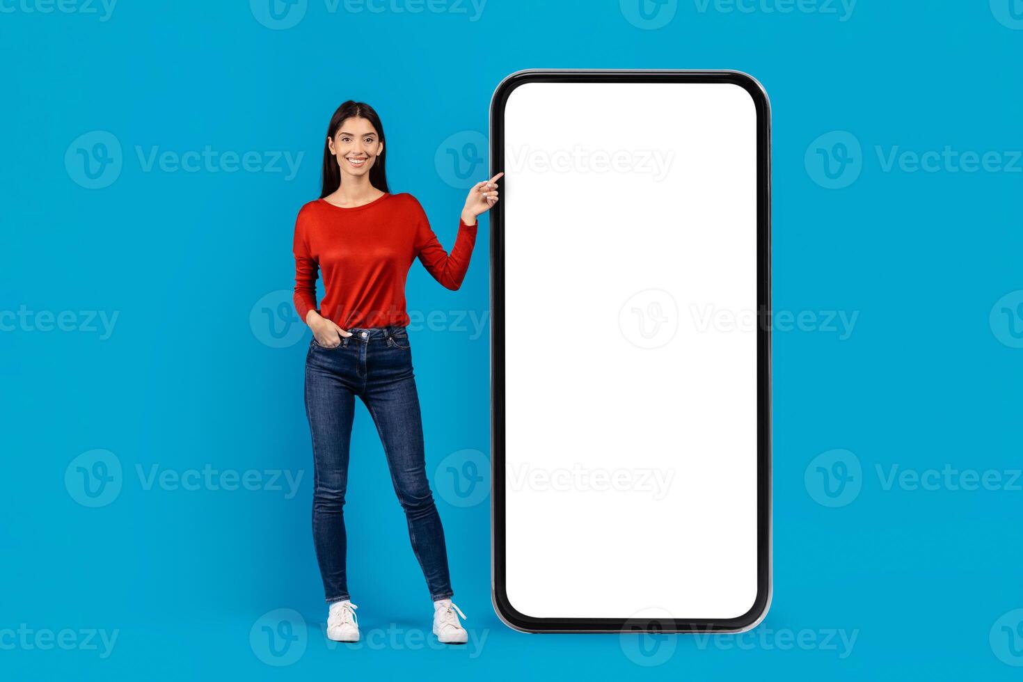 A smiling young woman dressed in a red top and blue jeans stands confidently next to an oversized smartphone model. She gestures towards the blank screen of the phone, mockup, copy space photo