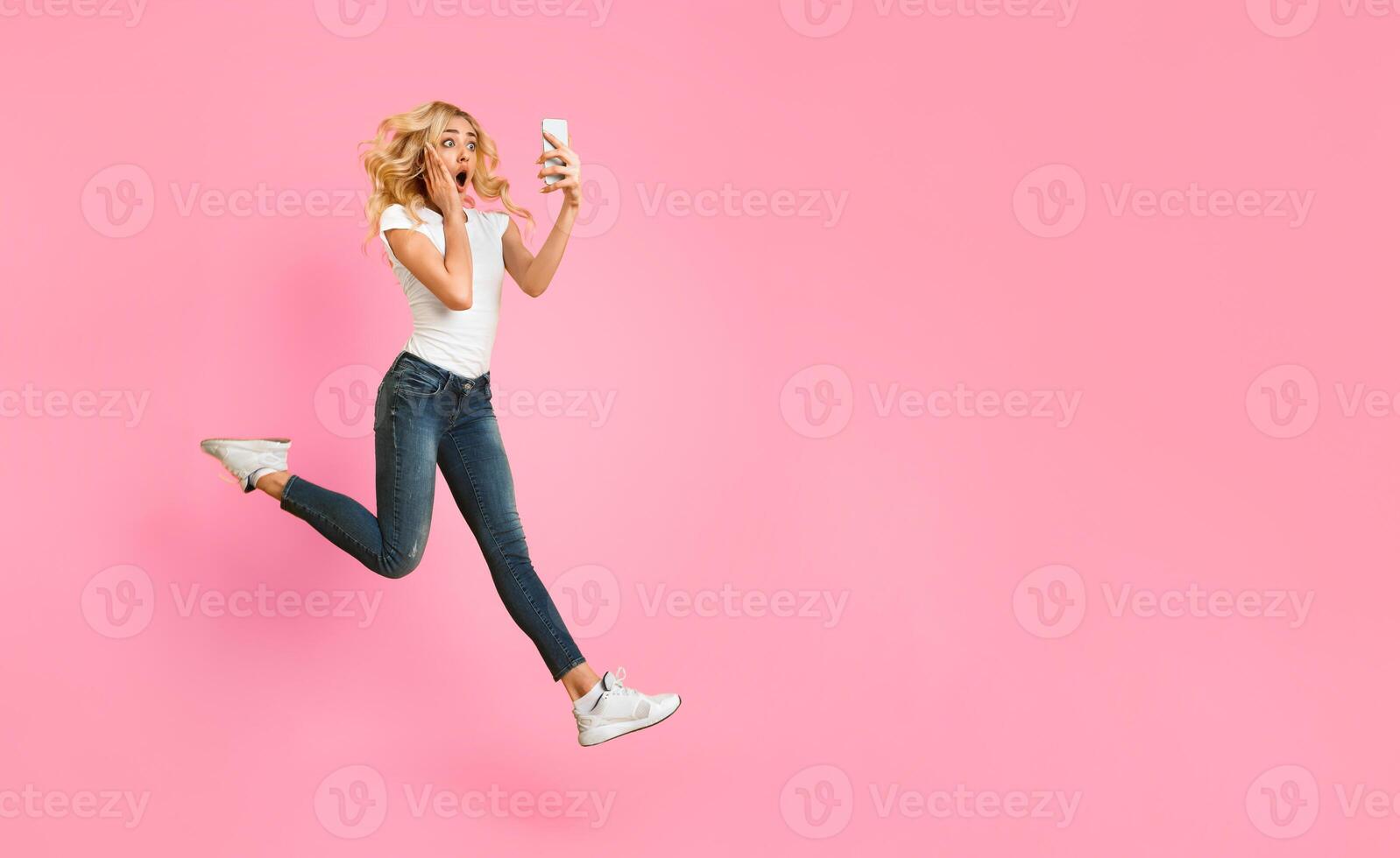 A young woman with blonde hair is jumping in the air with a surprised expression on her face. She is holding a white phone in her right hand and her left hand is on her cheek, copy space photo