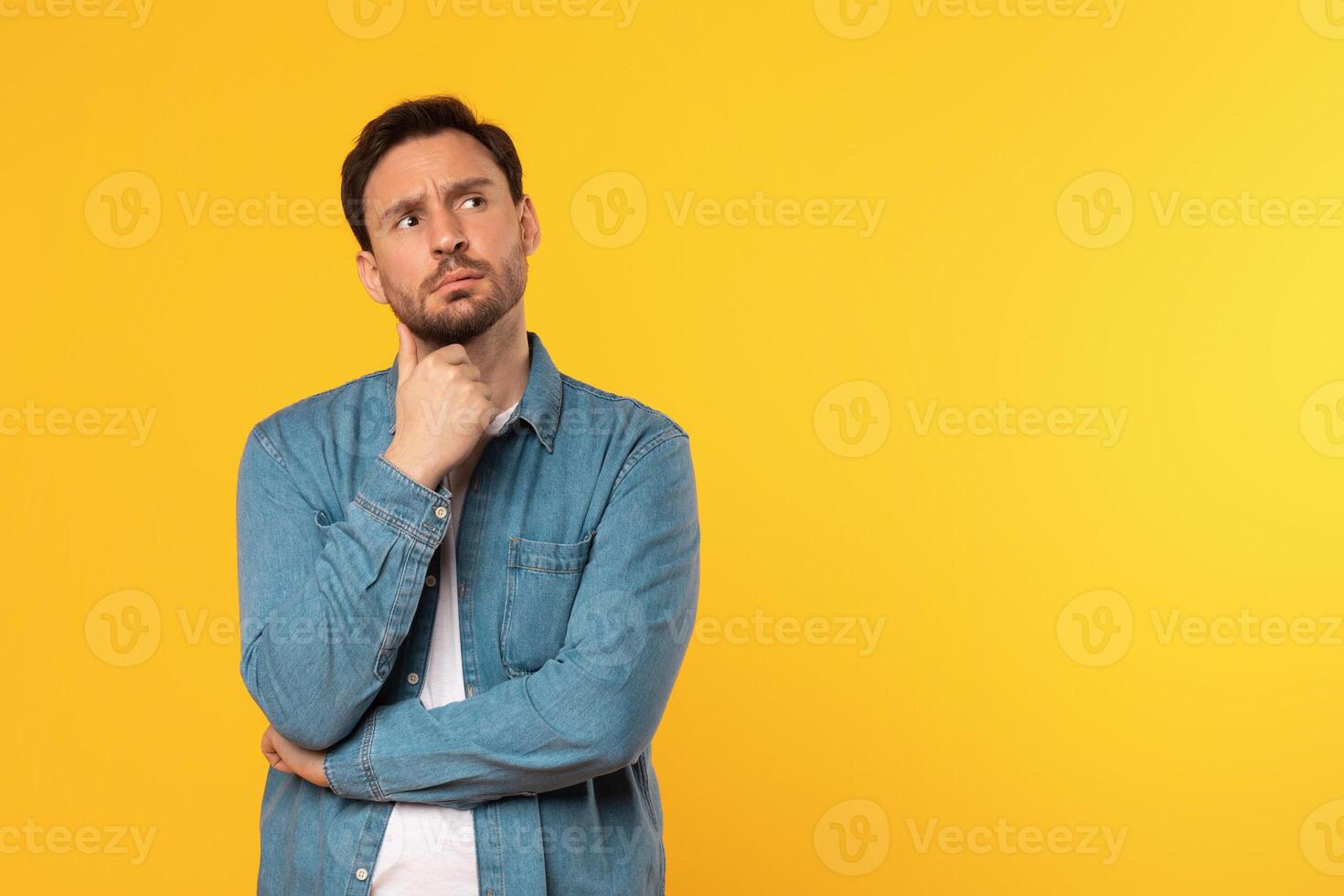 A man with a beard is wearing a blue denim shirt and looking at copy space while holding his chin with his hand. He is standing in front of a solid yellow background. photo