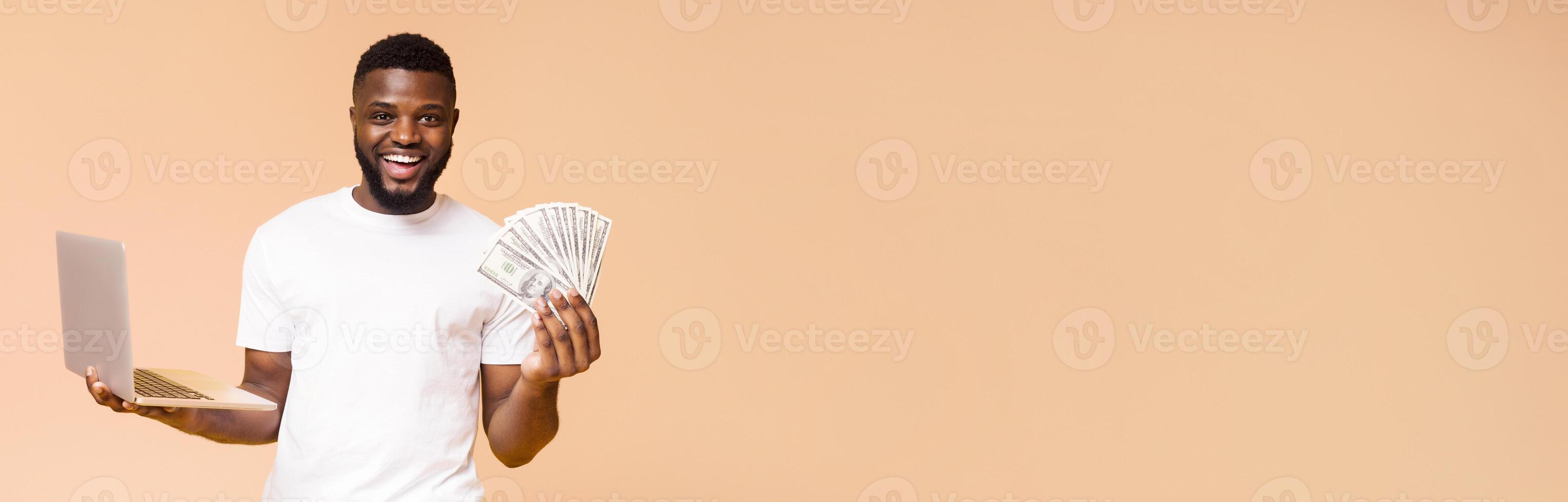 African American man is smiling and holding a laptop in one hand and a wad of cash in the other hand. He is standing in front of a solid beige background, copy space photo