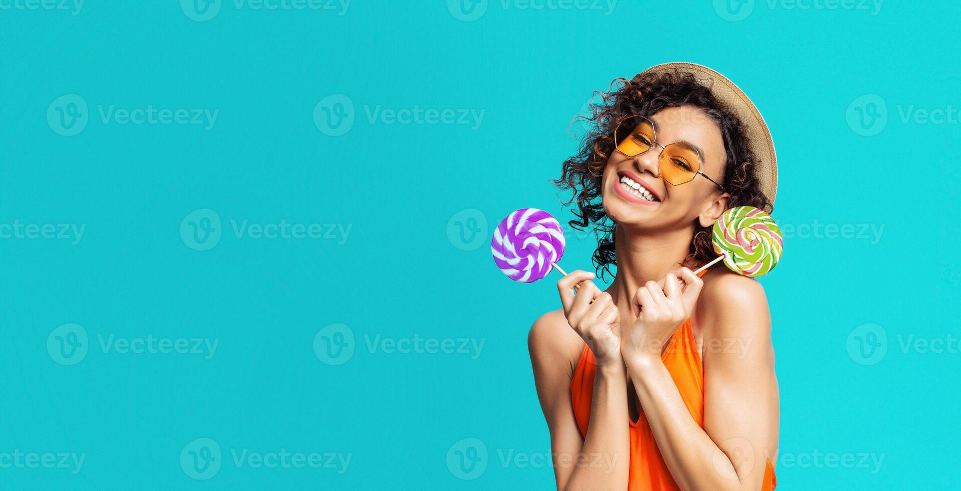 Black young woman with curly hair wearing sunglasses and a straw hat. She is smiling and holding two lollipops in her hands. The background is a bright blue color, copy space photo