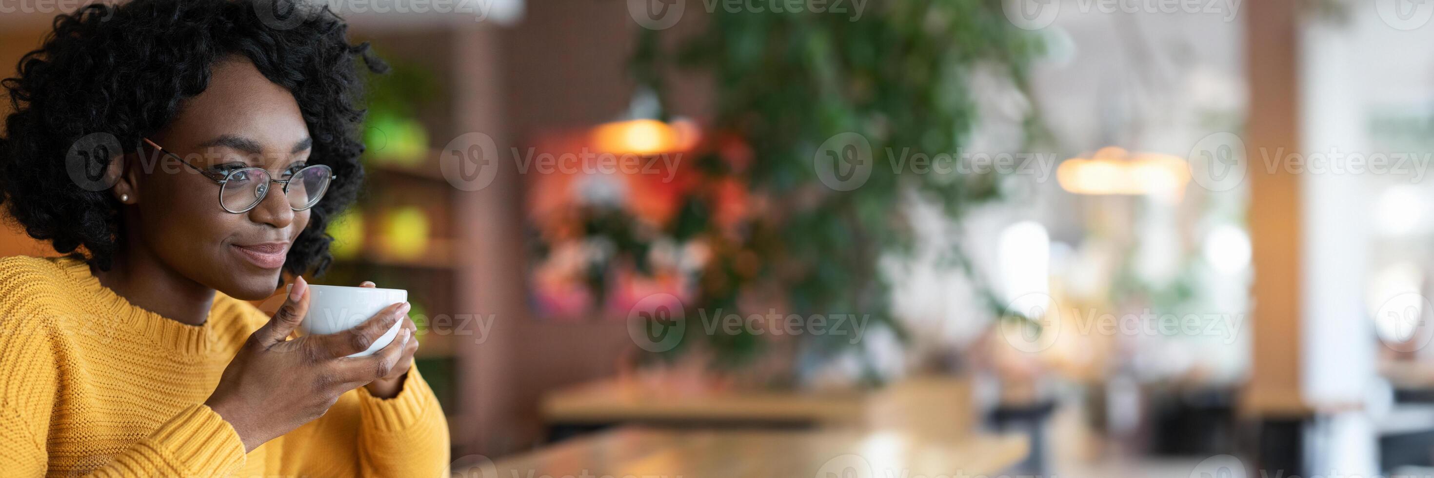 Dreamy young black lady in glasses enjoying cup of coffee in cafe, thinking about future opportunities, close up, copy space photo