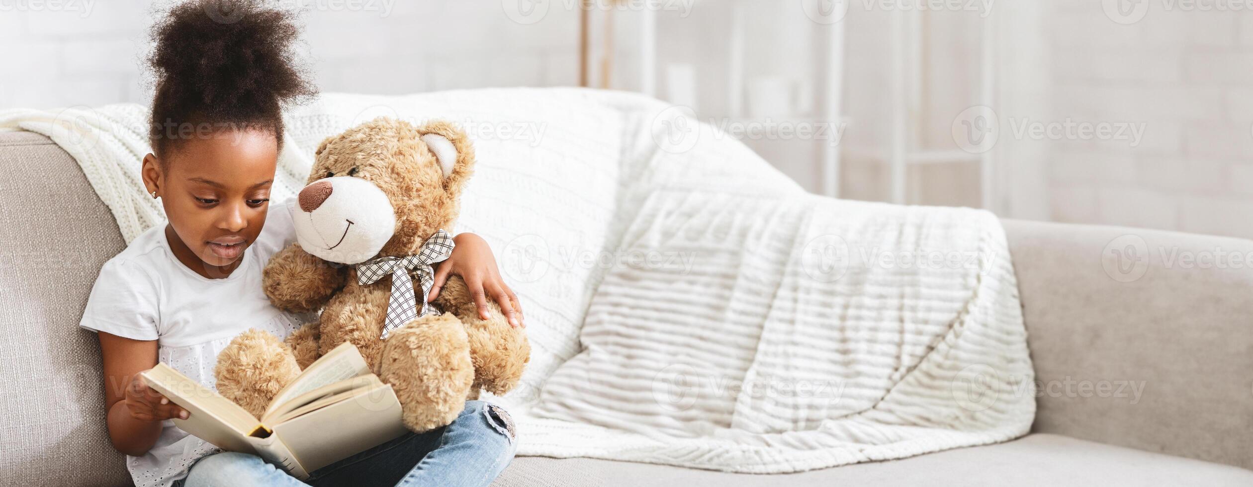 A young black child sits comfortably on a couch, deeply engaged in reading a book while holding a soft teddy bear. The atmosphere is warm and inviting, showcasing a moment of joy, copy space photo