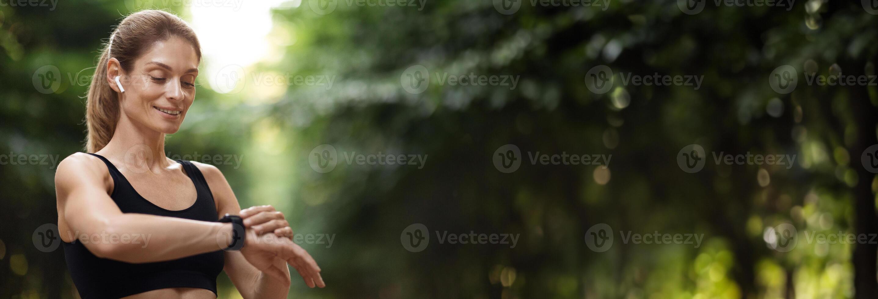 Closeup of happy pretty young sporty woman standing at public park, setting fitness tracker, long-haired millennial lady jogging outdoors, panorama with copy space. Technologies and sport photo