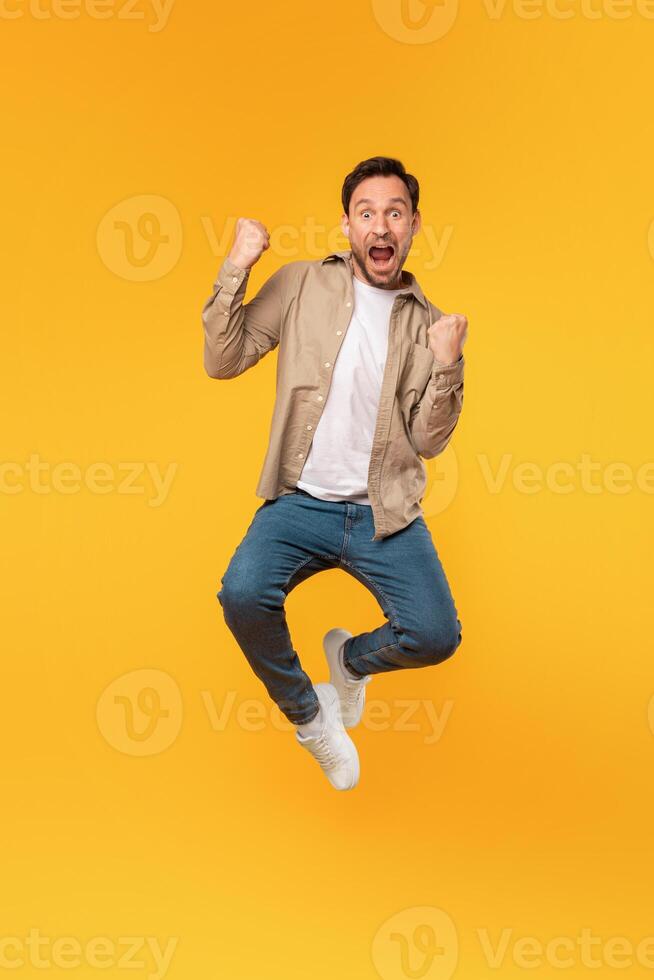A man with a beard and casual clothing leaps into the air, his fists clenched and mouth open in a shout of exhilaration against a yellow backdrop that radiates a sense of energy and happiness. photo