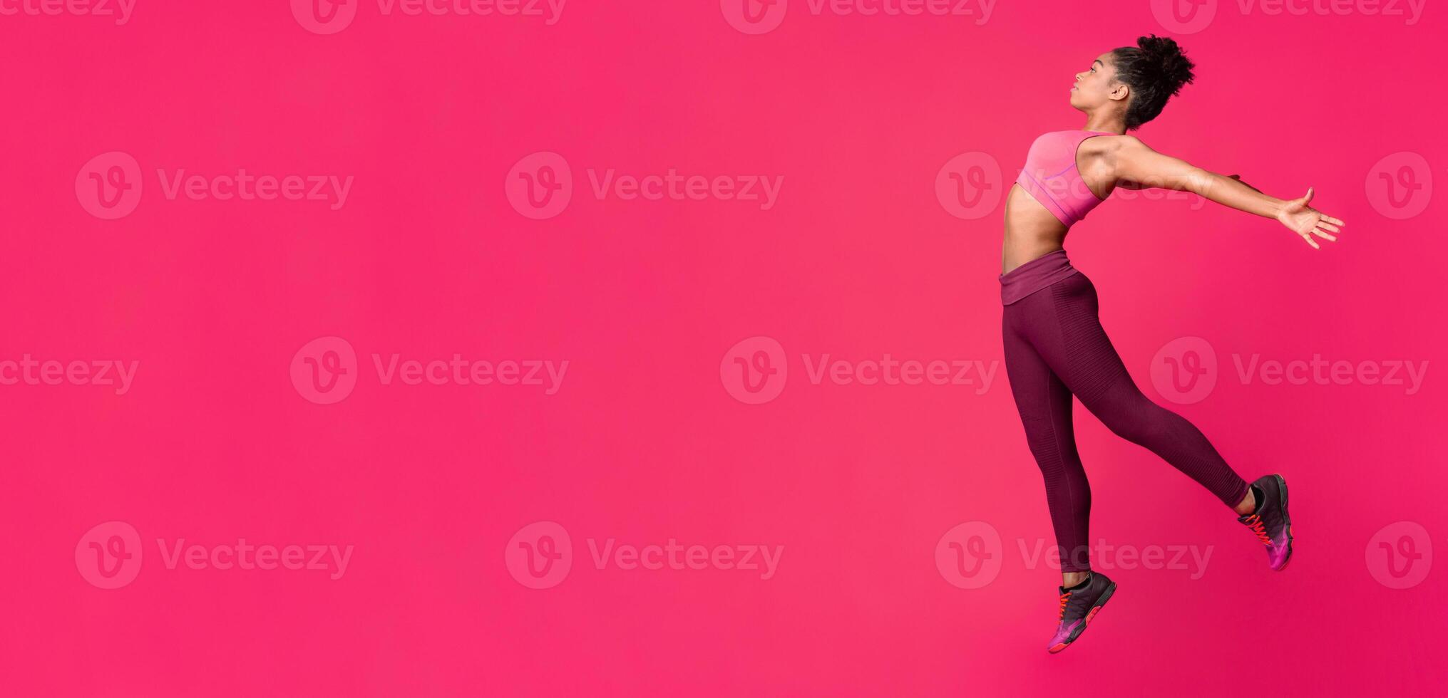 Flexible athletic black girl jumping up, stretching her body before training over red background, side view photo
