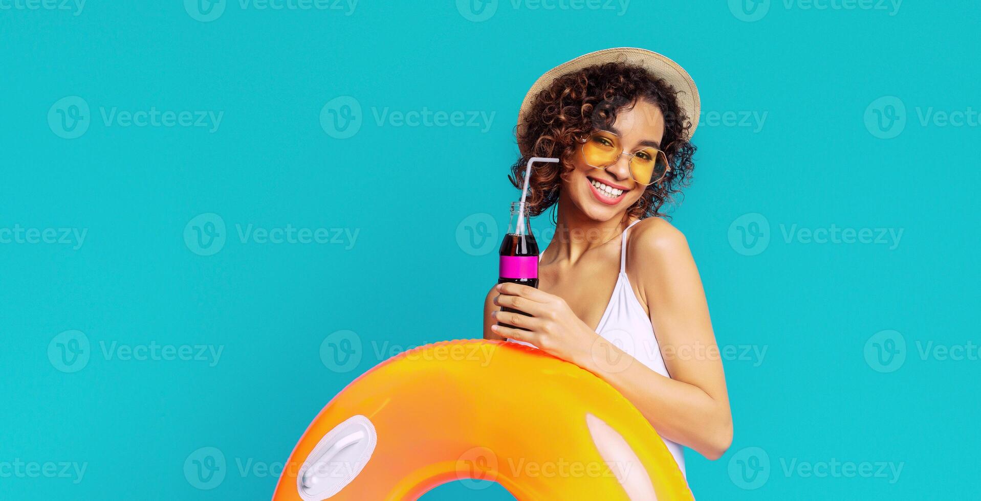 Black woman with curly hair smiles brightly as she poses in front of a blue backdrop. She is holding a soda with a straw and an inflatable ring, suggesting a day of fun and relaxation in the sun. photo