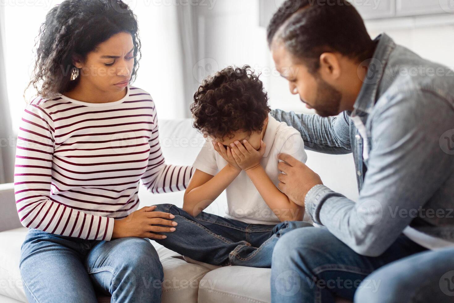 African American man and woman are seated on a couch, accompanied by a child. Parents comforting their upset crying child, home interior photo