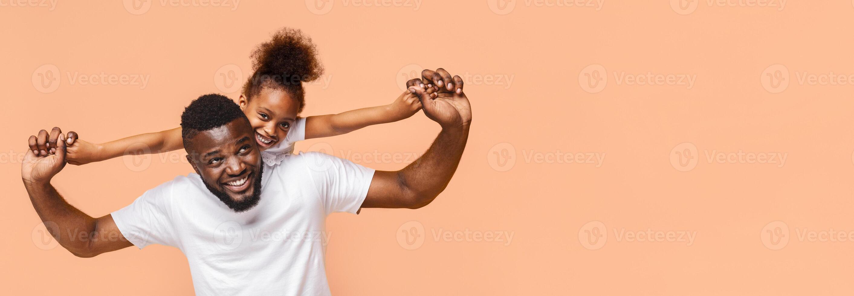 A joyful black father and daughter are playing together in a studio setting. The father is holding the daughter on his back, and they are both smiling and laughing, panorama with copy space photo