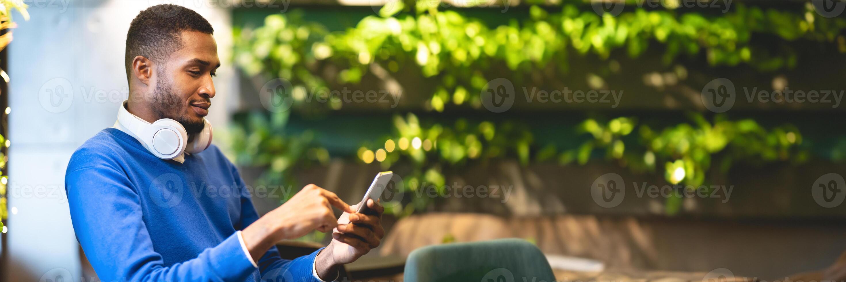 Technology Concept. Side view of young african american student in headphones using smartphone sitting at modern cafe, panorama with copy space photo